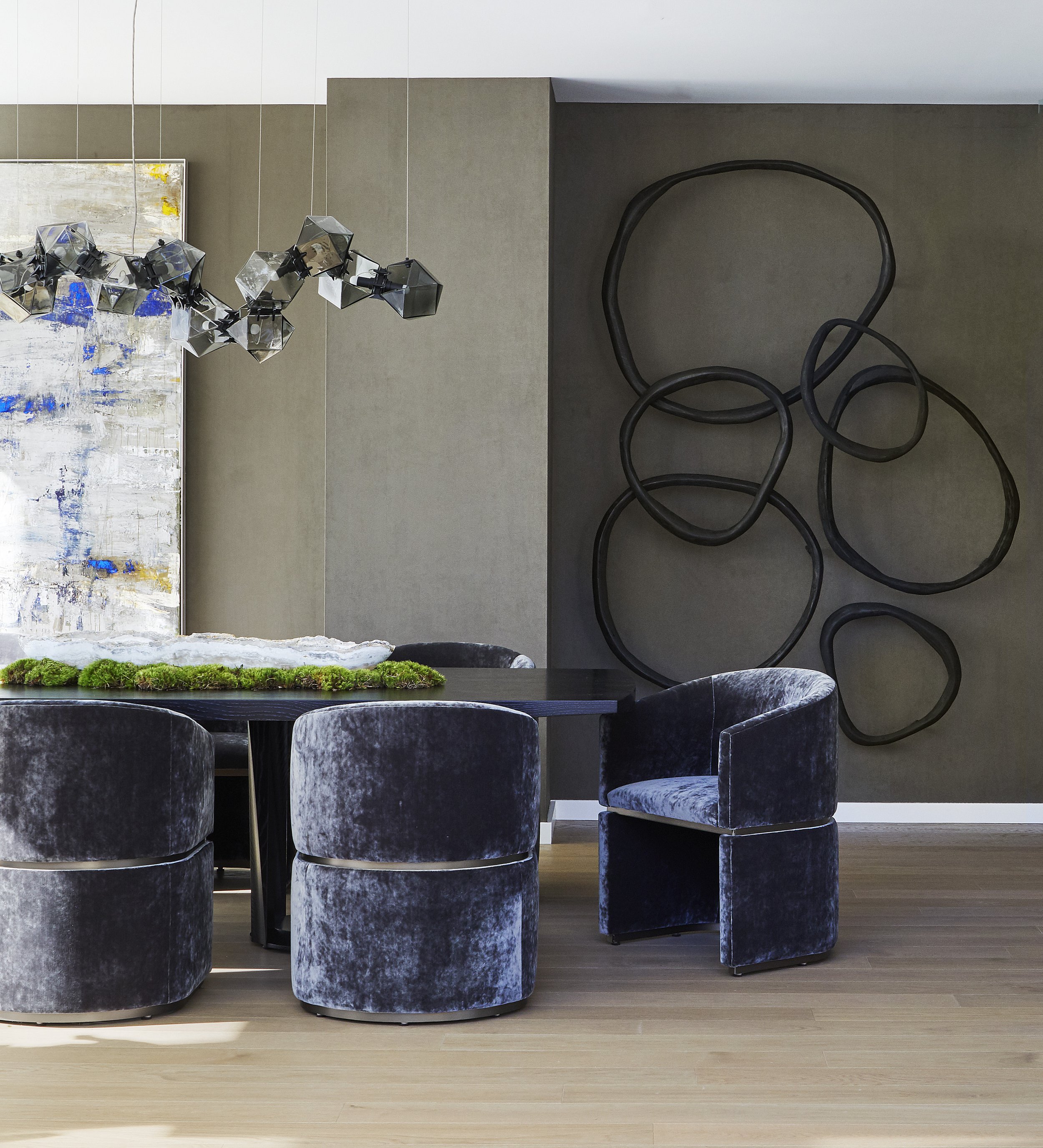 Modern dining room with a black marble table, velvet upholstered chairs, a large abstract painting, a geometric chrome chandelier, and a sculpture on the wall.
