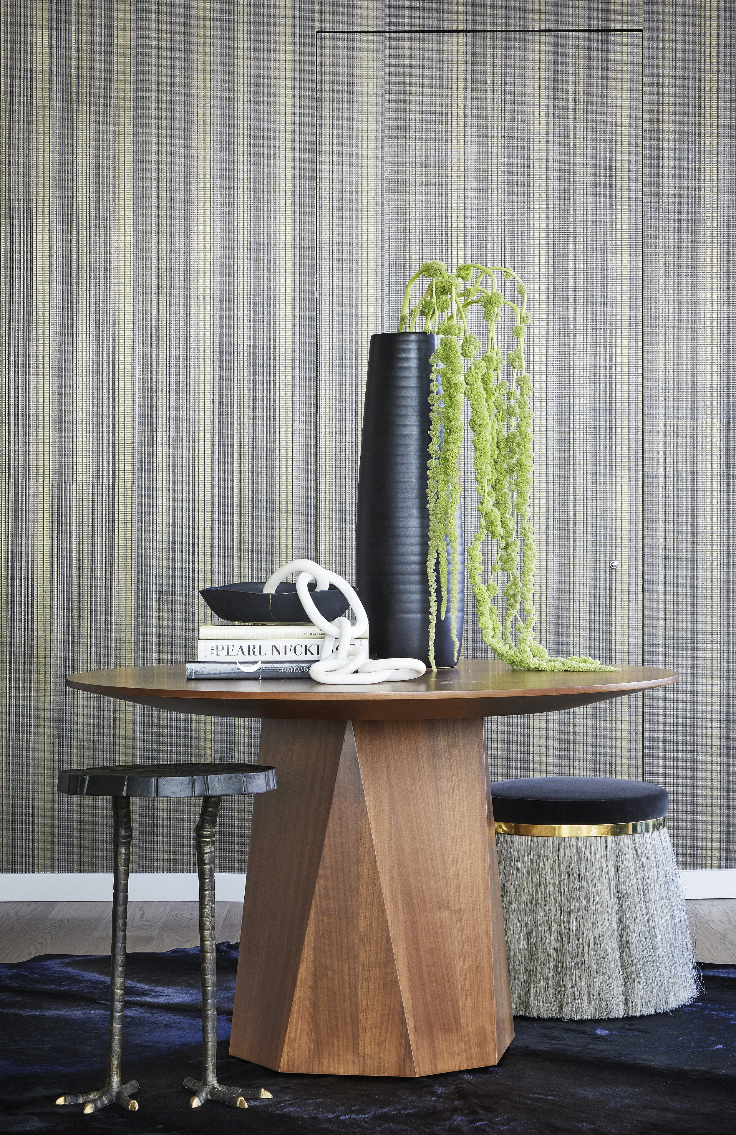 A wooden round dining table with modern and vintage-style chairs, decorated with a black vase containing green hanging plants and a white decorative chain, set against textured gray wallpaper.