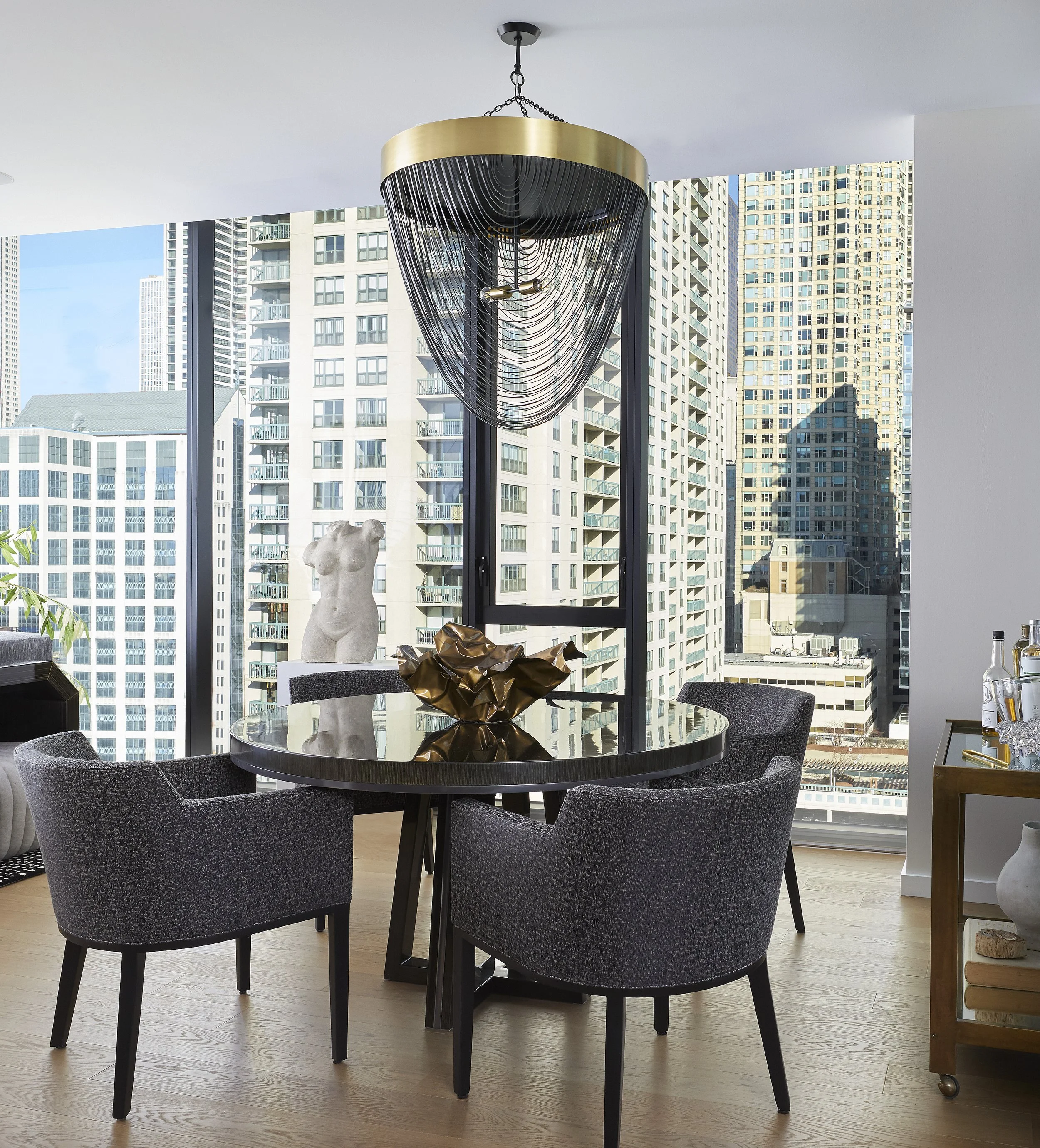 A modern dining room with a round dark wood table, six upholstered dark gray chairs around it, and a decorative centerpiece shaped like a crumpled gold metallic bowl. In the background, large windows reveal a cityscape with tall buildings, some with 
