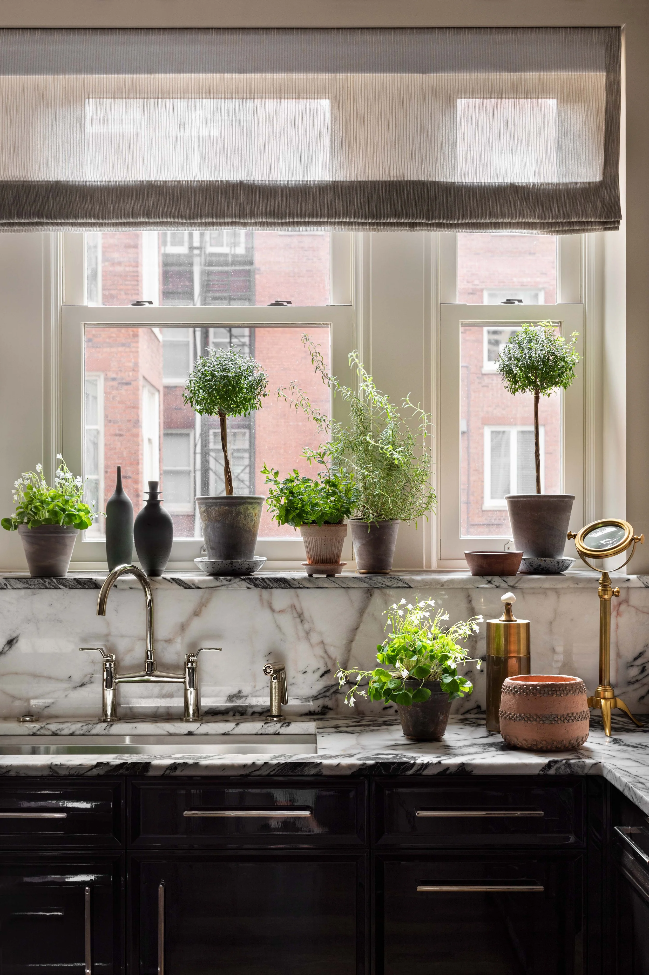 Kitchen window ledge with various green houseplants in pots, a marble countertop sink with silver fixtures, and a window with a semi-transparent window valance and city buildings outside.