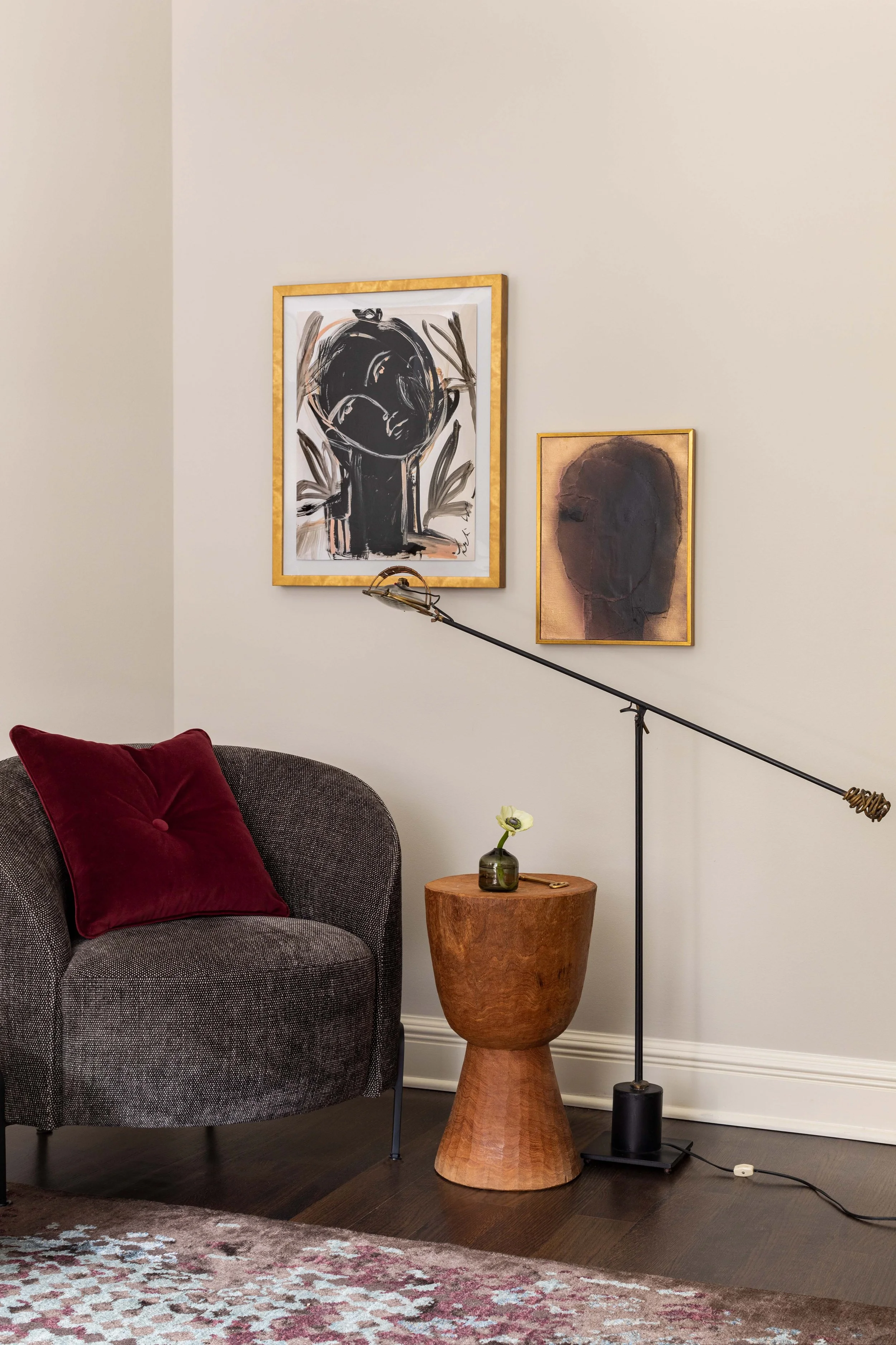A living room corner with a dark gray armchair with a red velvet pillow, a wooden side table with a small green vase and a flower, two abstract framed artworks on a beige wall, a black floor lamp with a long adjustable arm, and a patterned area rug o