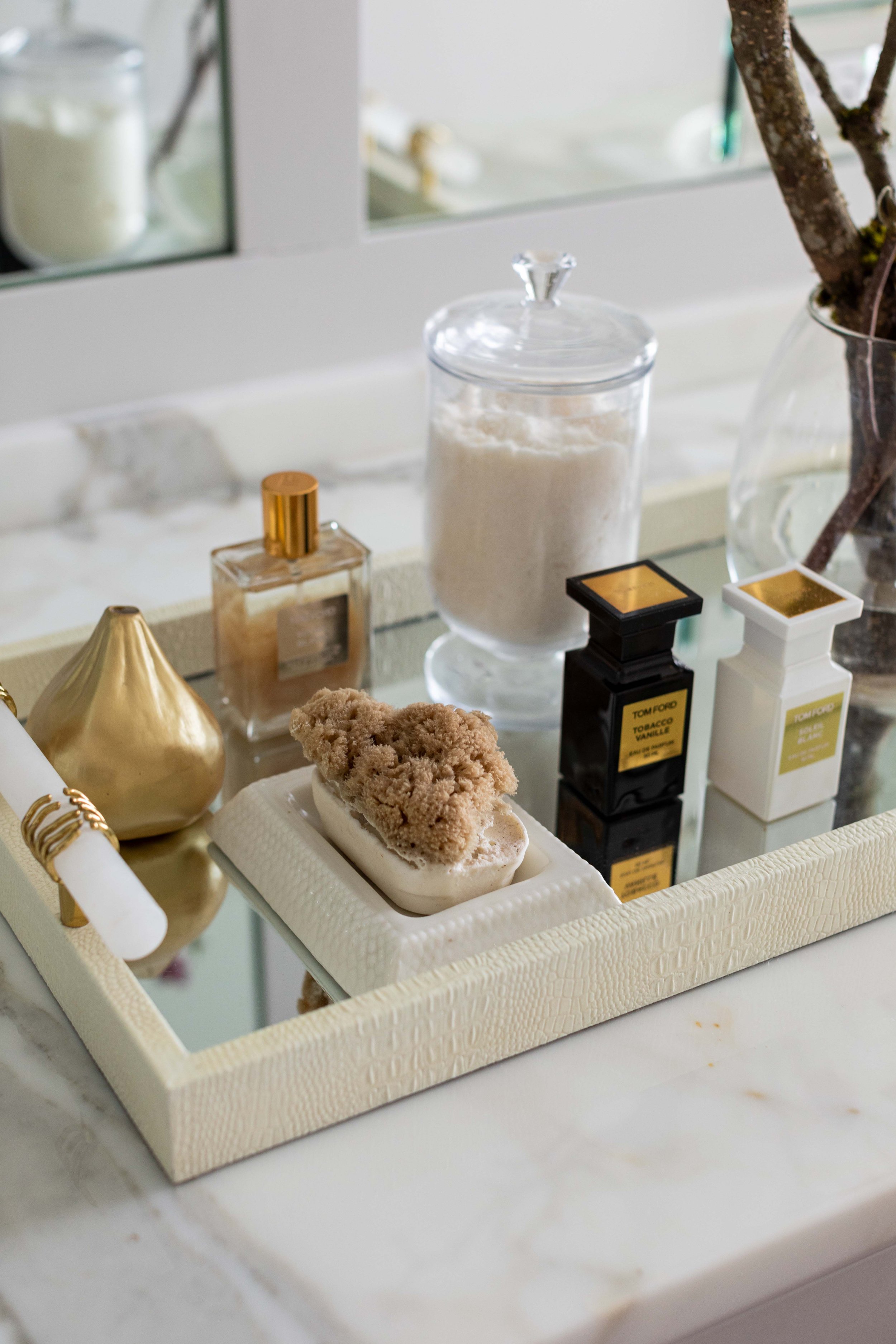 A tray with various luxury perfume bottles and a soap with a loofah on top, placed on a marble surface with a mirror and a glass vase with branches in the background.