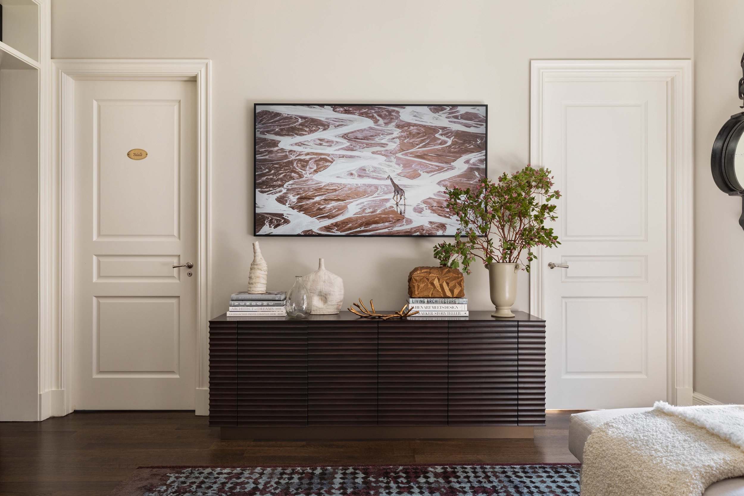 Living room interior featuring a framed aerial photograph of a river delta, a dark wooden sideboard with decorative vases, a stack of books, a potted plant, and a patterned rug.