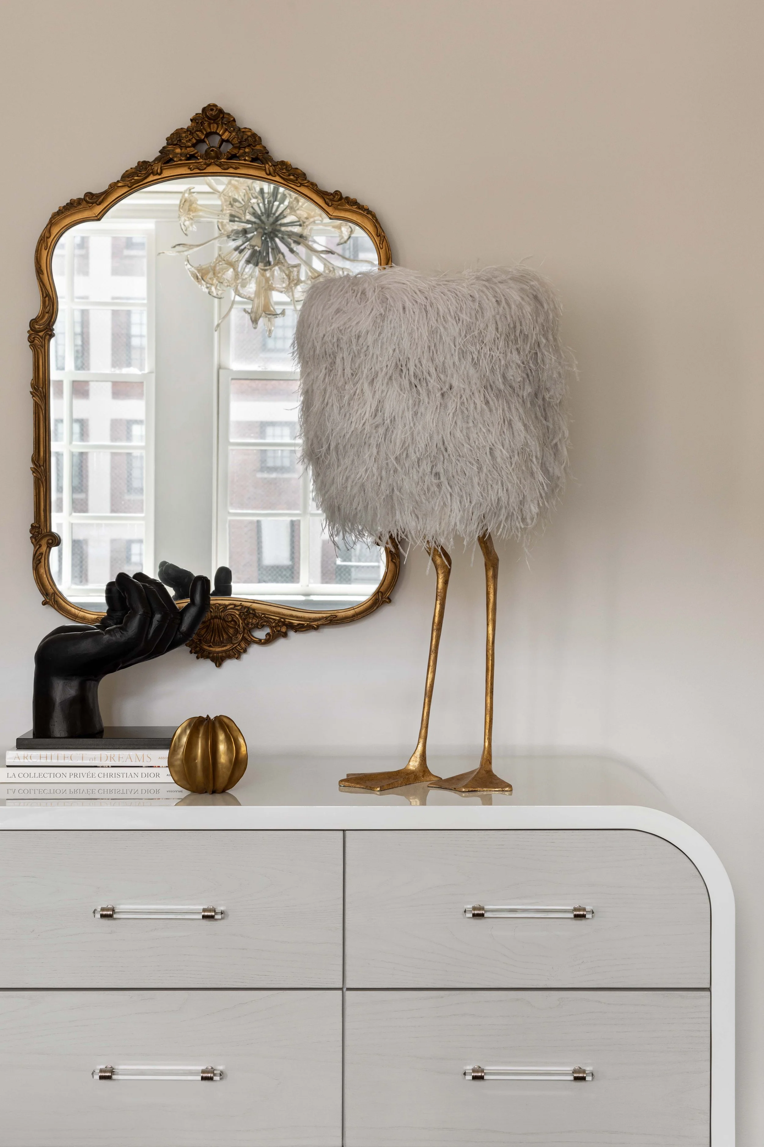 A white dresser with silver handles, decorated with a black hand sculpture, a small gold pumpkin, a stack of books, a feathered white bird figure with long legs, a gold-framed mirror, and a chandelier with floral details reflected in the mirror.