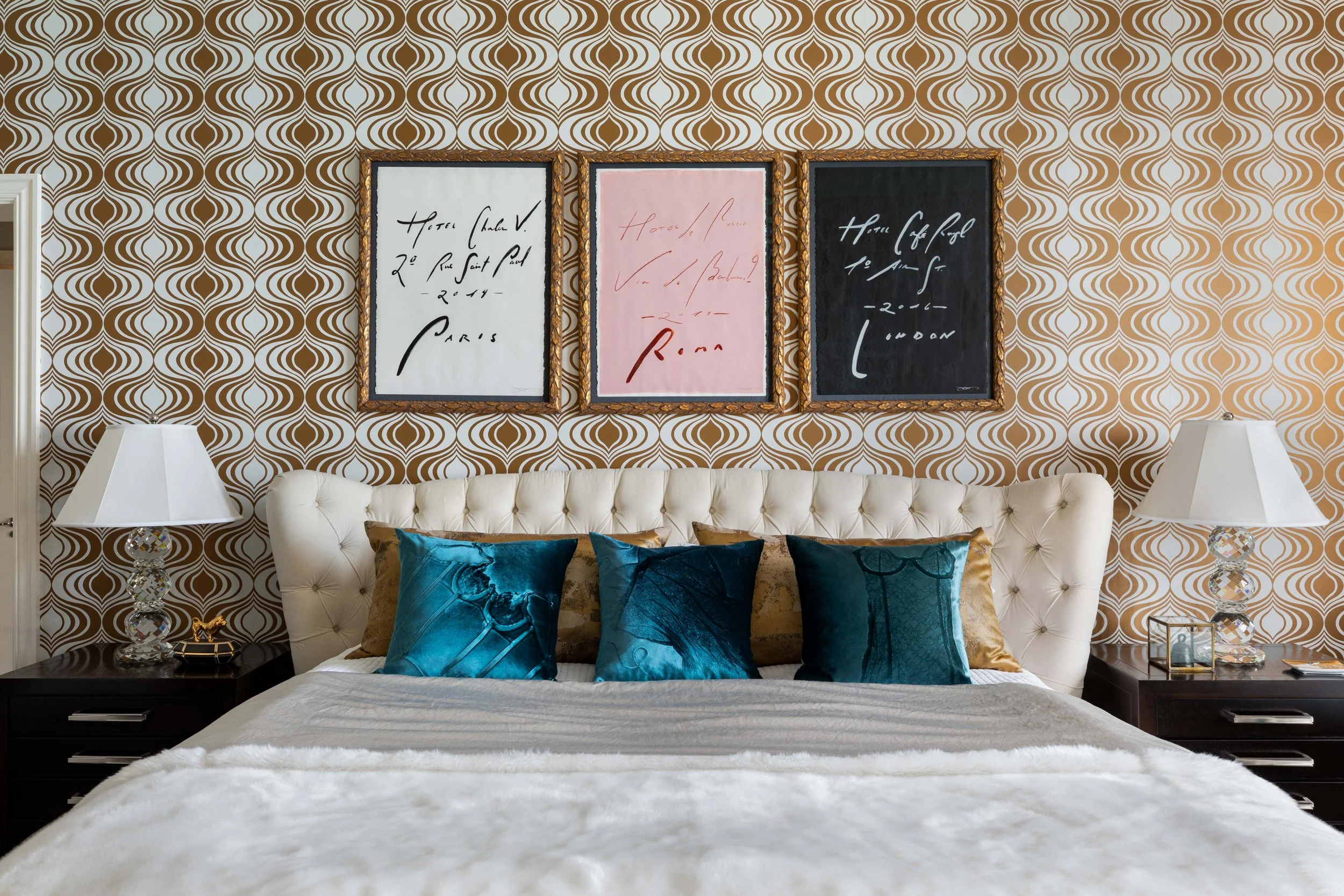 A bedroom with a tufted white headboard, decorated with blue and gold pillows, a black nightstand with a glass lamp, and three framed art pieces with handwritten text on a patterned brown and white wallpaper background.