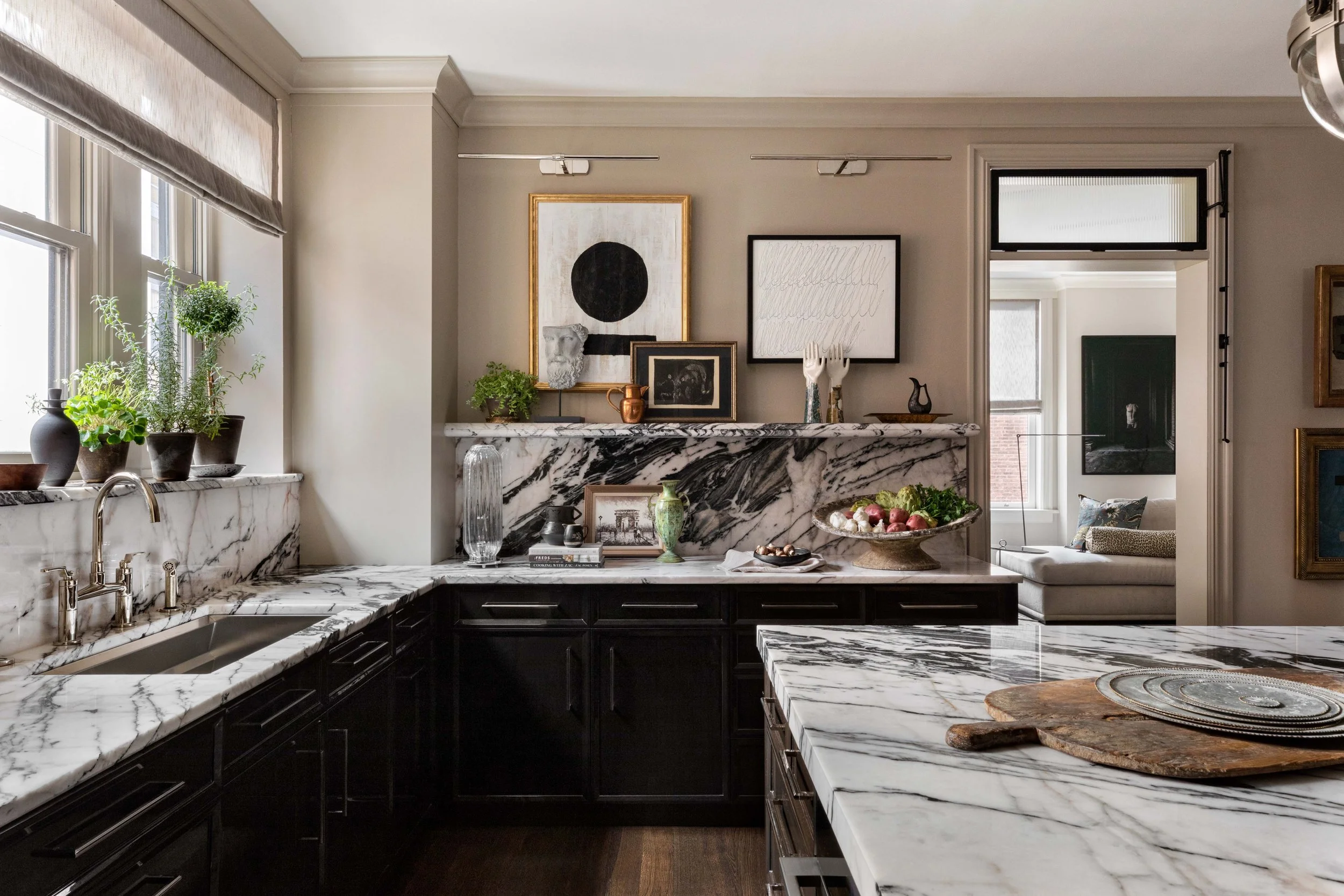 A kitchen with marble countertops and backsplash, black cabinetry, and various decorative items on the counter, including potted plants, framed art, a vase, and a bowl of apples. There are windows with beige blinds and framed artwork on the wall.