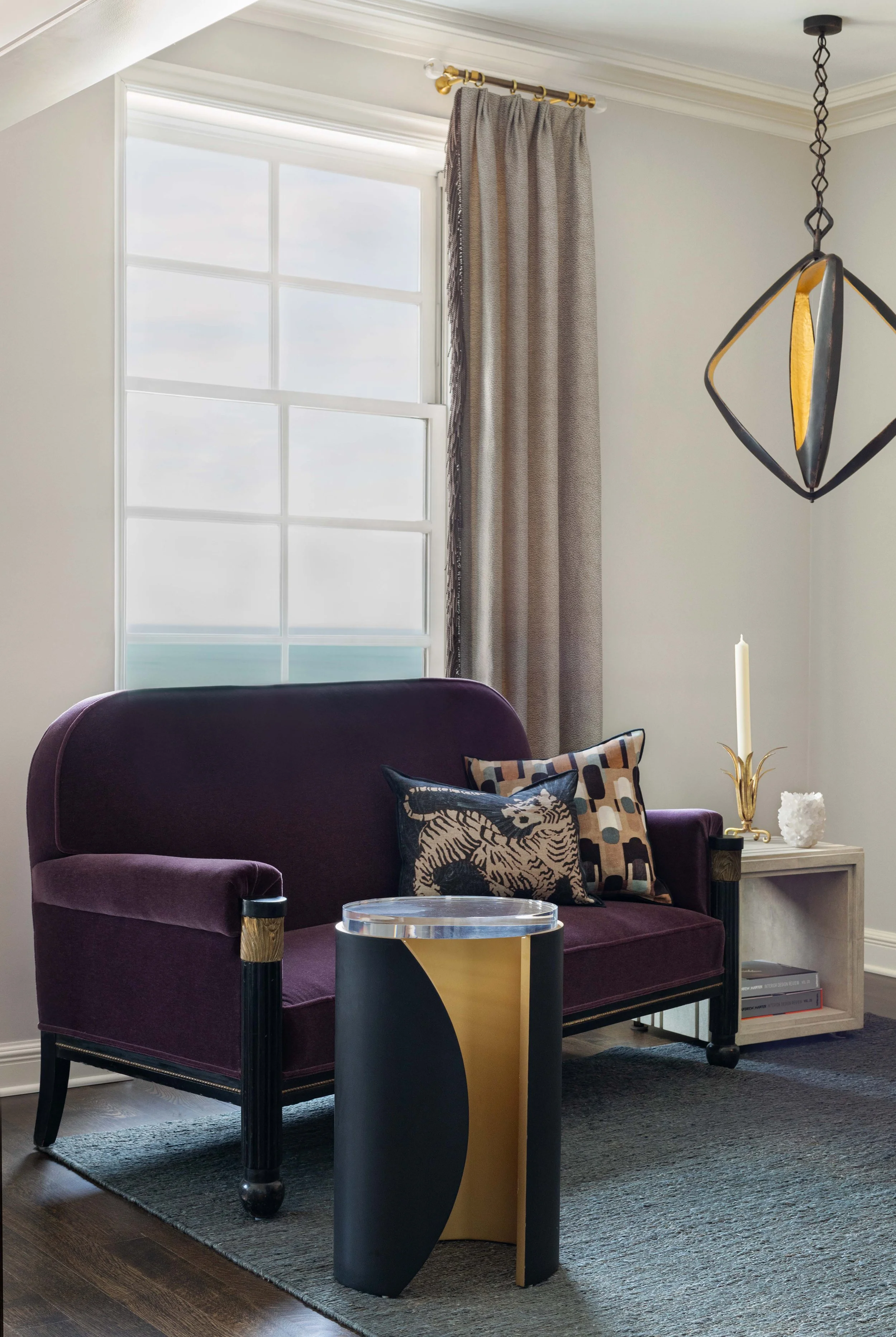 Living room corner with a purple velvet sofa, patterned throw pillows, a white side table holding a white candle in a gold holder, black and gold coffee table, a modern hanging light fixture, and a large window with beige curtains.