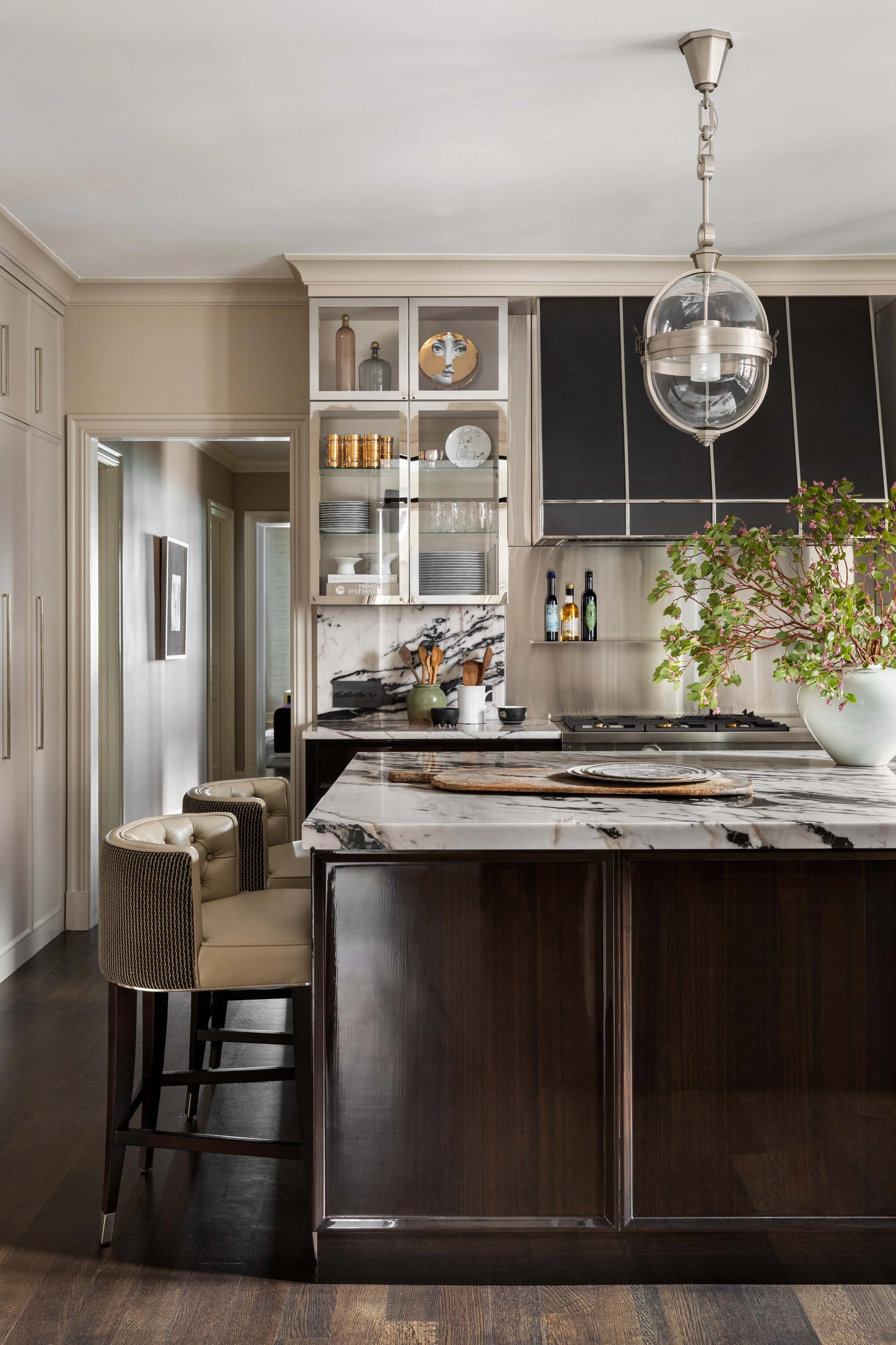 Kitchen with a marble island, dark wood cabinets, a pendant light, open shelves with dishes and bottles, and potted plant.