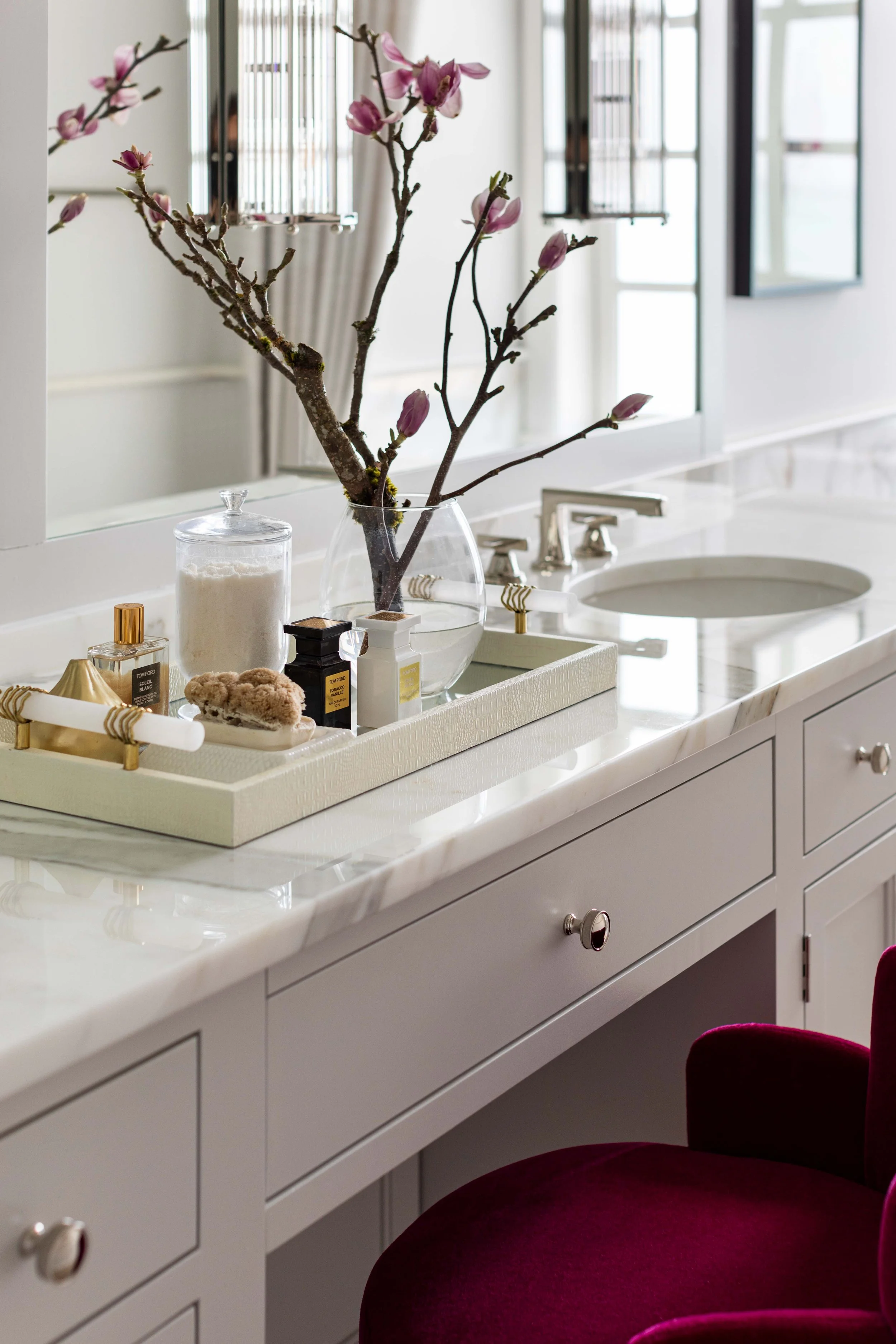 A bathroom vanity with a white marble countertop, a large mirror, and various toiletries including perfumes, a jar of cotton balls, and a vase with pink magnolia branches. There is a burgundy velvet chair at the edge of the image.
