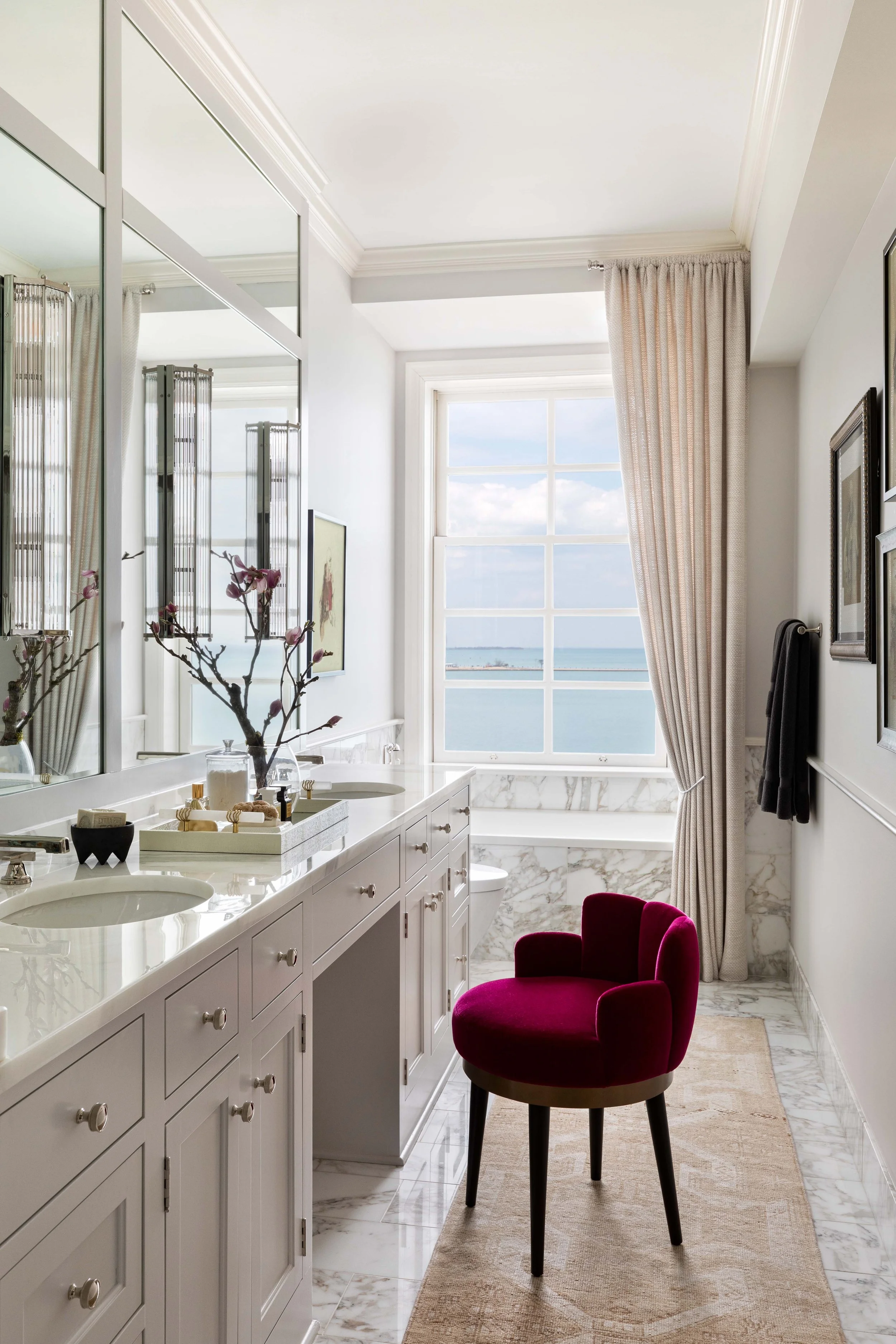 A bright bathroom with white cabinetry and marble countertops. There is a window with a view of the ocean and sky, decorated with beige curtains. A red velvet chair is placed on a beige rug, and a branch with pink flowers is on the counter.