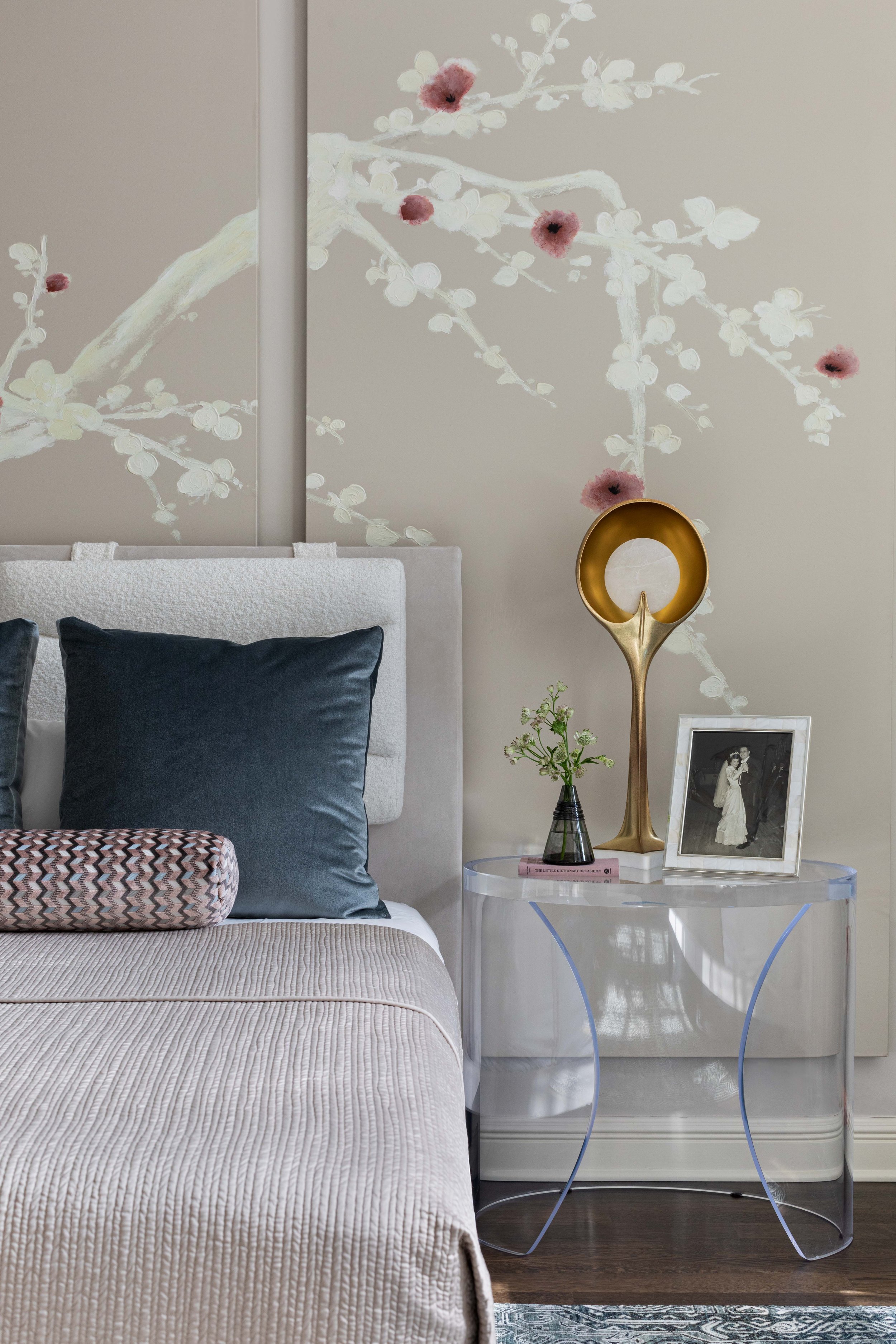 A bedroom with a beige bed, dark blue pillows, and a decorative pillow with a zigzag pattern. A white side table holds a framed black-and-white photo, a green plant in a black vase, a pink book, and an abstract gold sculpture. The wall behind feature