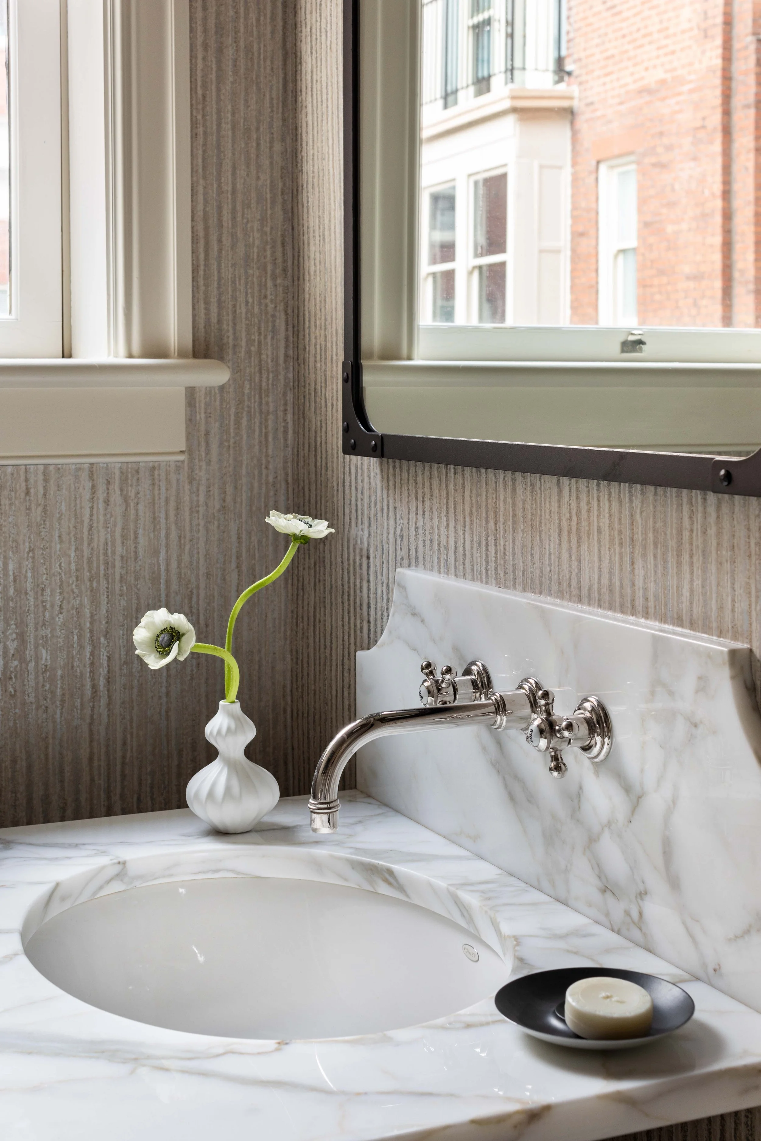 A bathroom vanity area featuring a round white marble sink, a silver faucet, and a soap dish with a candle. There is a white vase with white flowers on the marble countertop. Behind, a window shows neighboring brick and stone buildings. The wall has 