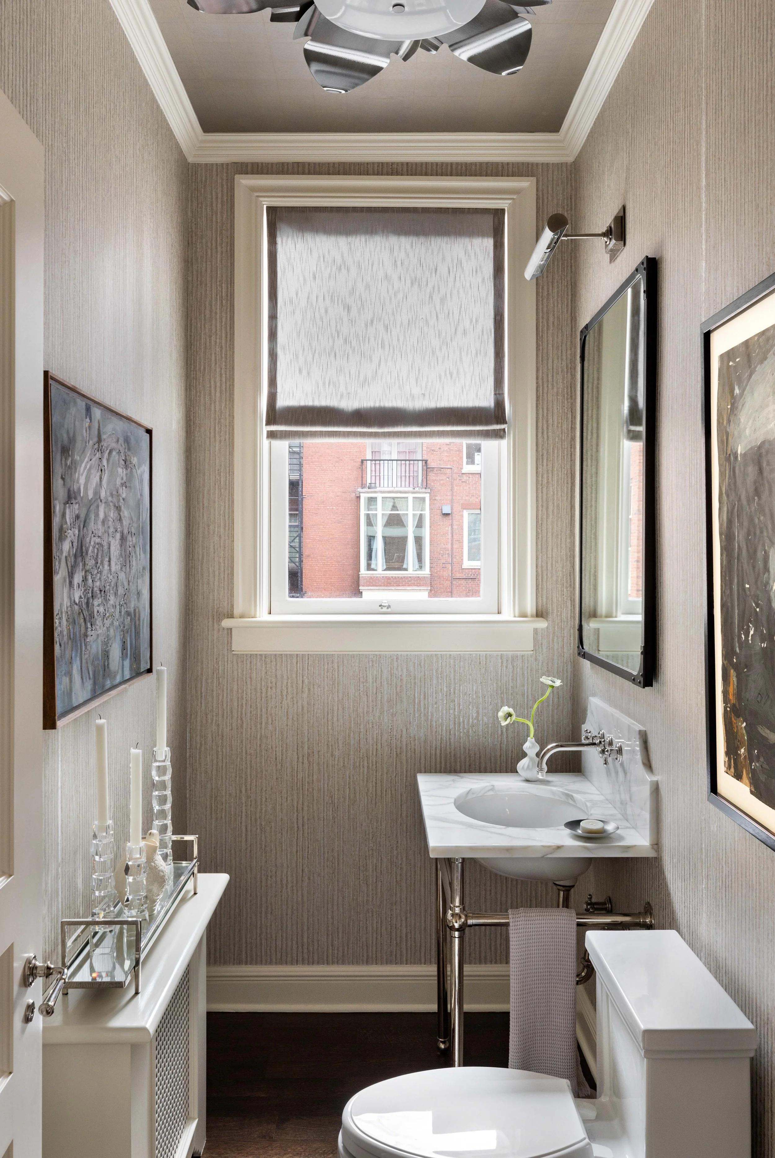 A small, elegant bathroom with vertical striped wallpaper, a window with a gray roller shade, a marble sink with a small flower vase, a mirror, and a white toilet, with a ceiling light fixture with reflective petals.