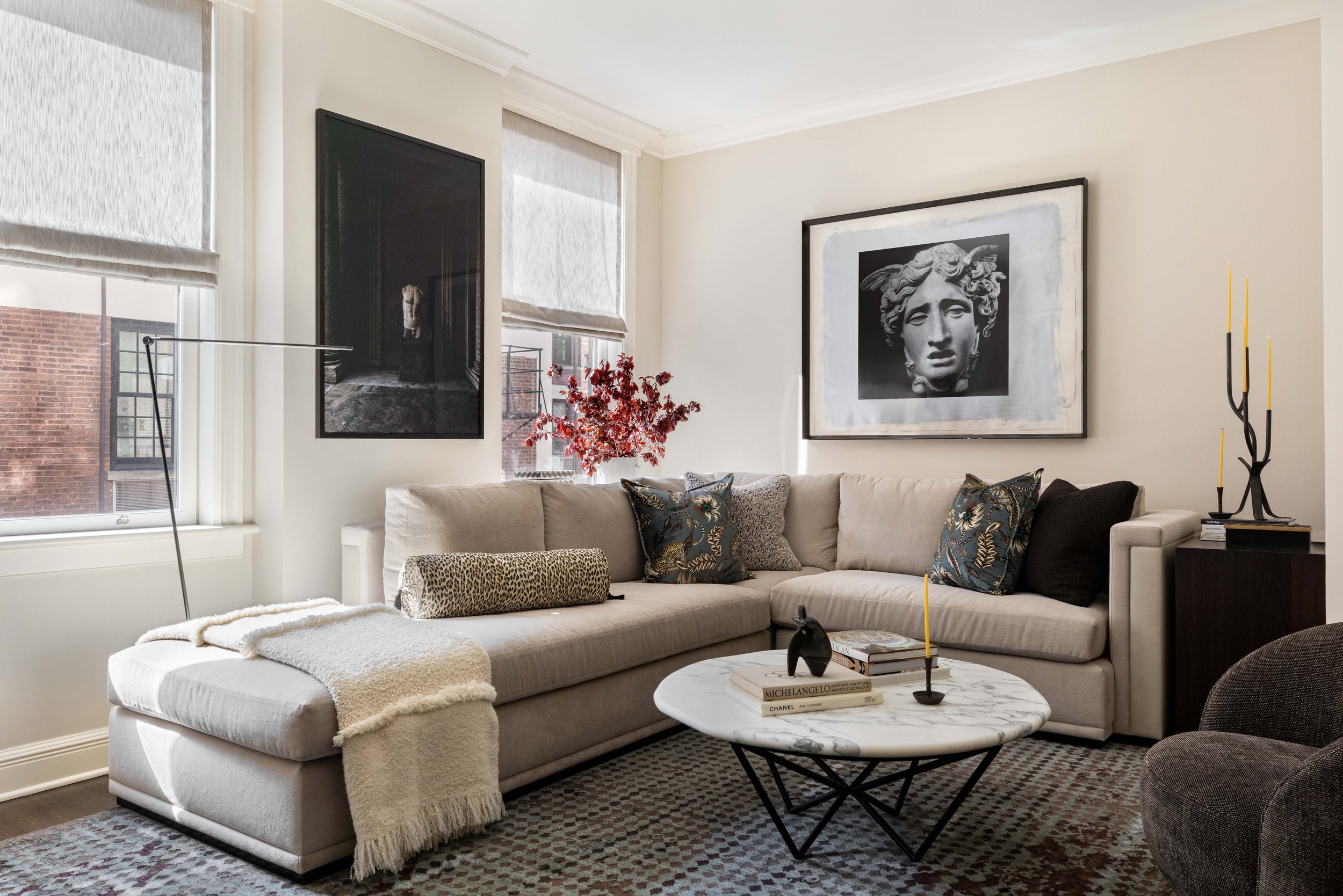 A living room with beige sectional sofa, decorative pillows, marble coffee table, wall art, large windows with shades, and a patterned rug.