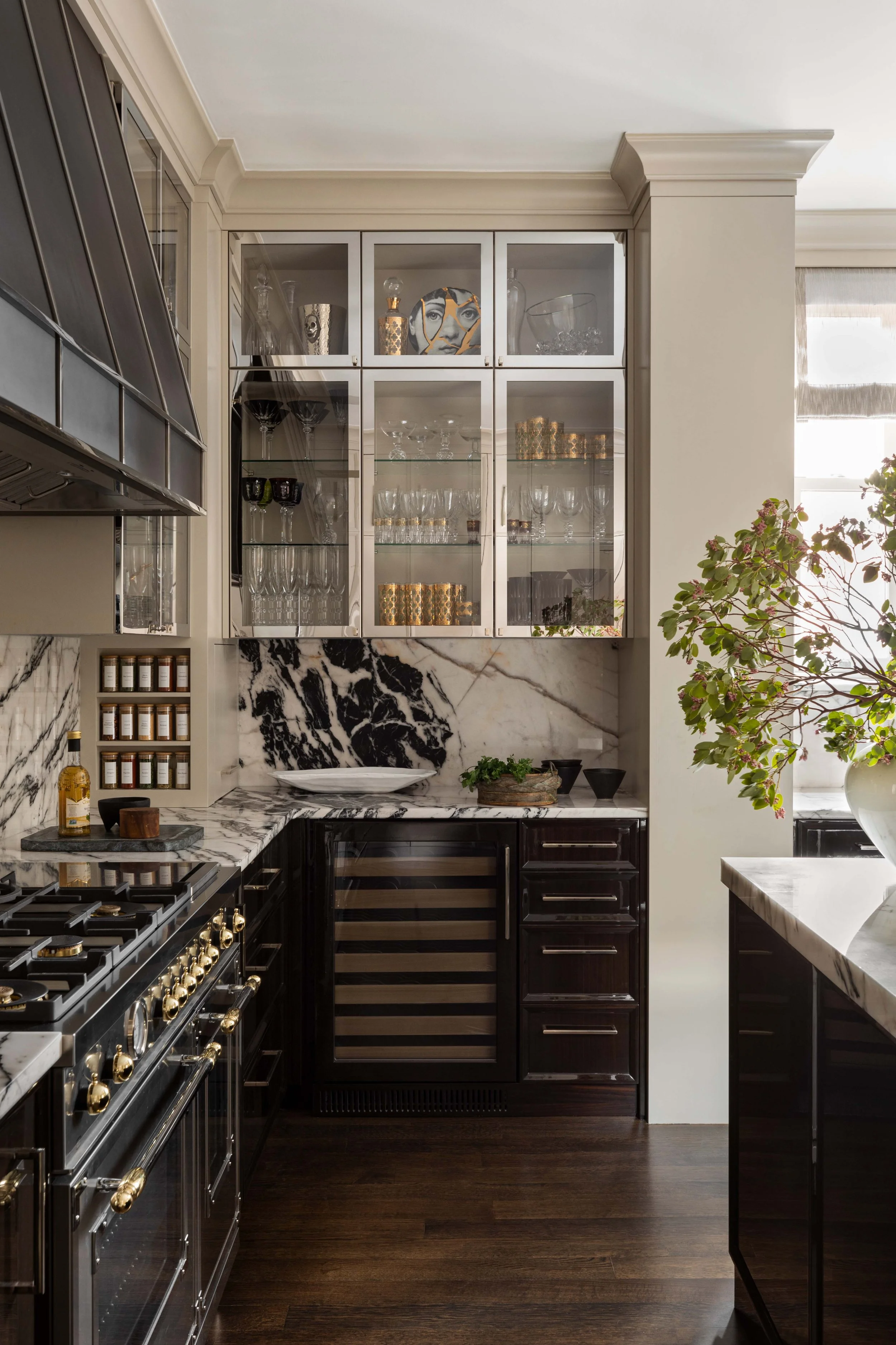 A kitchen with black cabinets, marble countertops and backsplash, glass cabinet filled with glassware, a wine cooler, and a small table with a potted plant and decorative bowls.