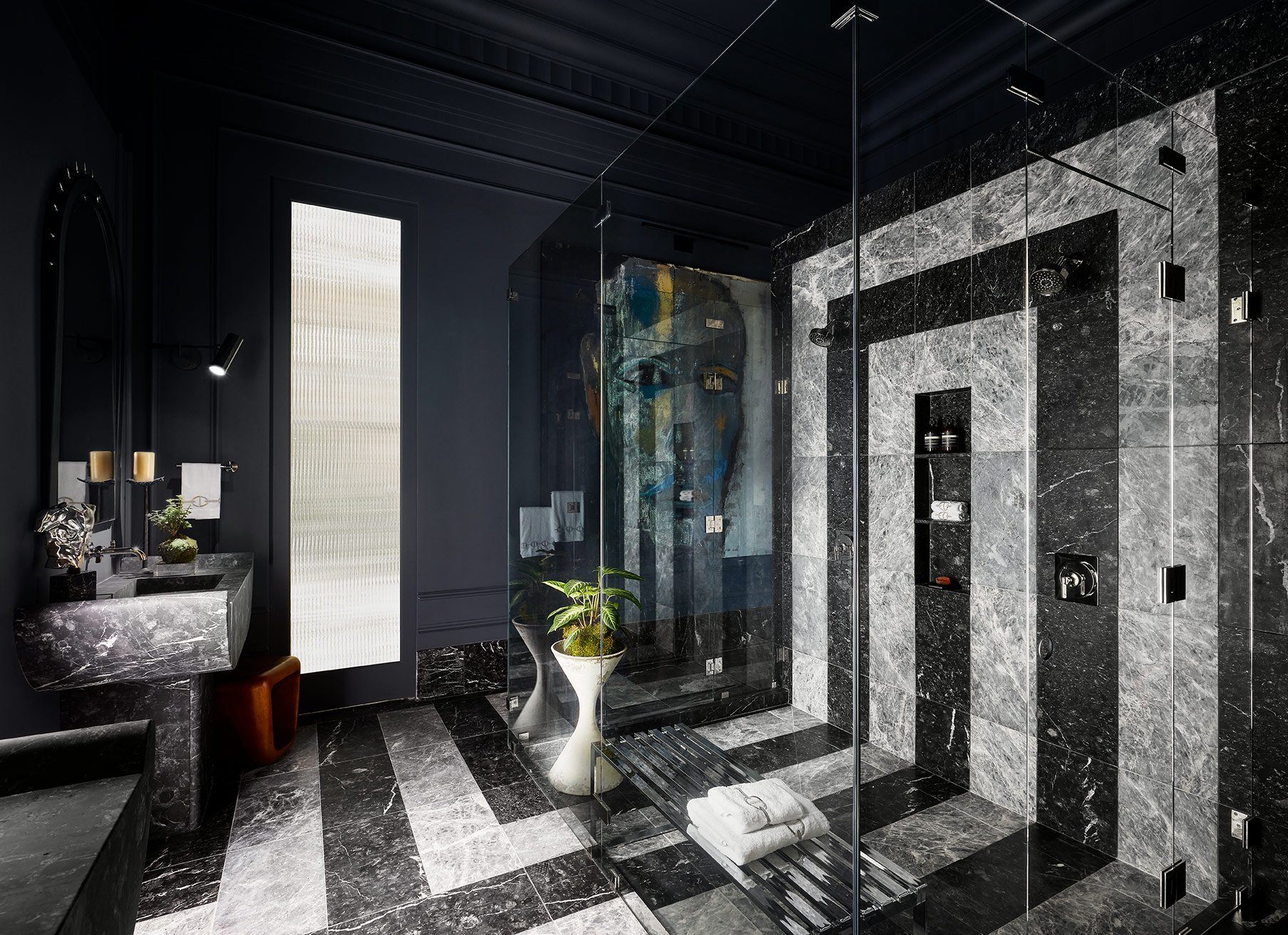 A modern bathroom with black and gray marble tiles, a glass shower enclosure, a floating vanity with a marble countertop, and black fixtures. There is a small potted plant and decorative accessories near the sink, and a textured light panel on the wall.