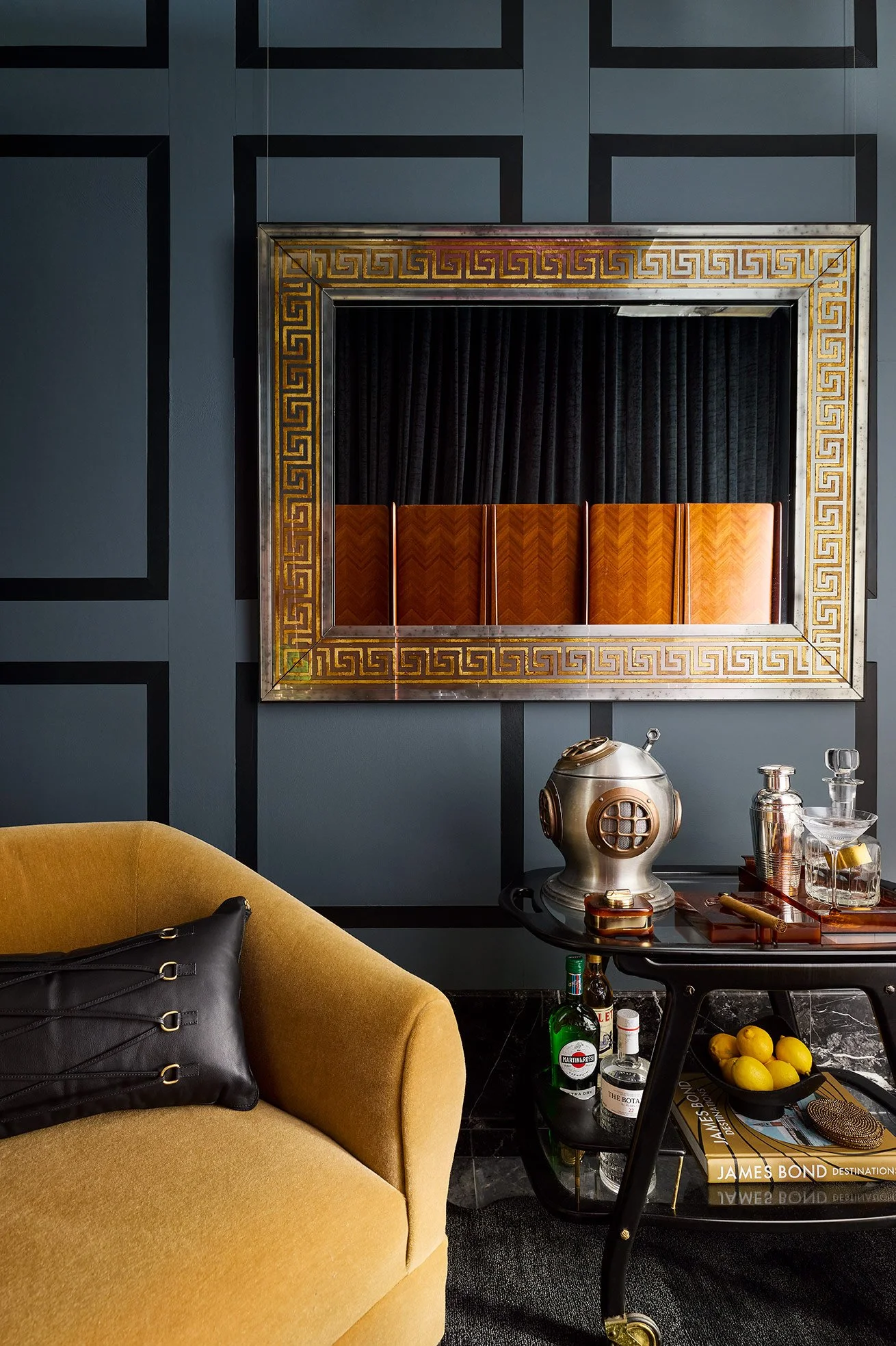 Interior room with a mustard yellow sofa, a black pillow, a small black coffee table holding bottles, lemons, and reading materials. A wall features a decorative mirror with a Greek key pattern, black curtains, and a dark blue paneled wall. Items on 