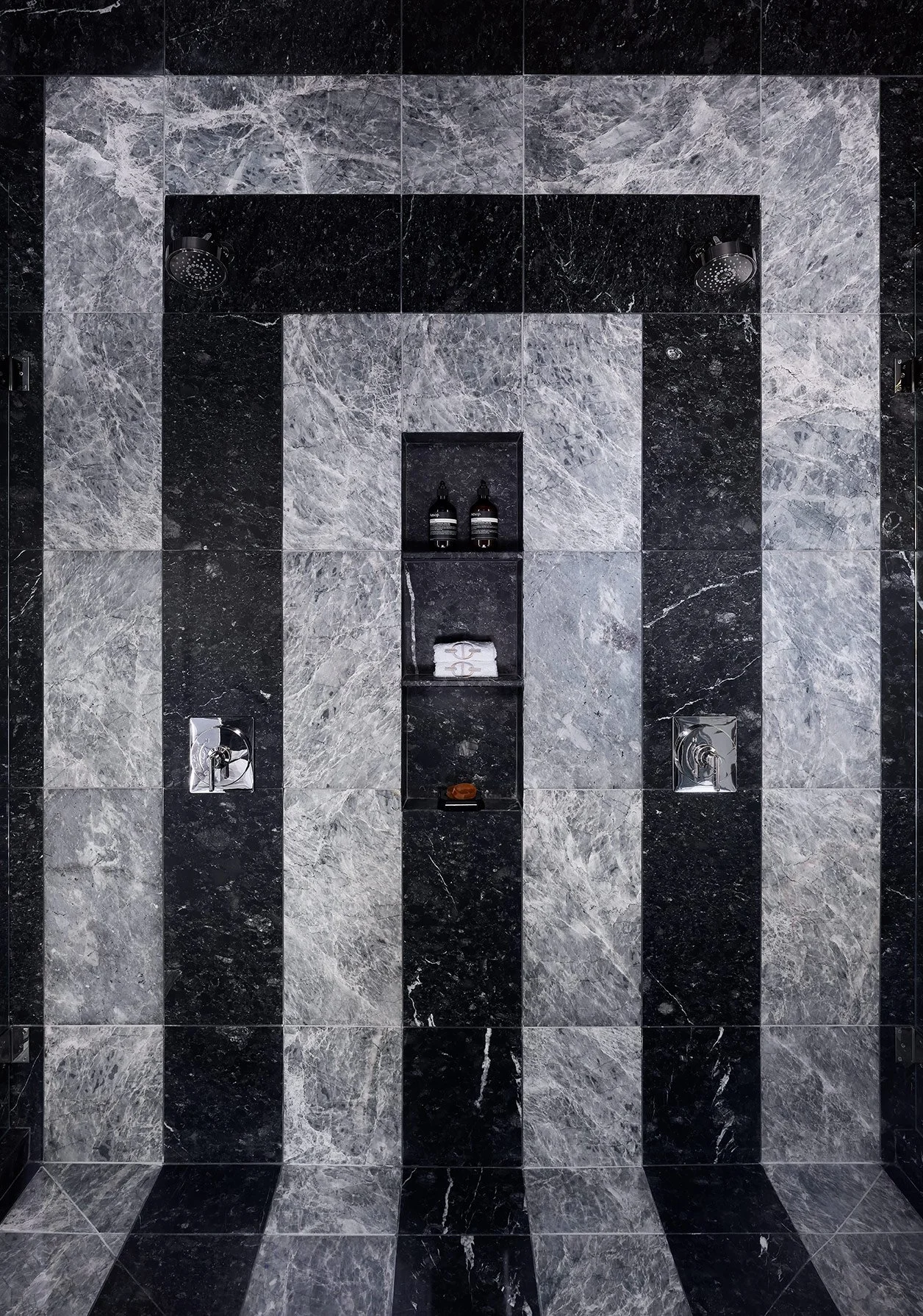 View of a modern showerspace with marble tile walls and floor, two showerheads, a central shelf with toiletries, and control valves.