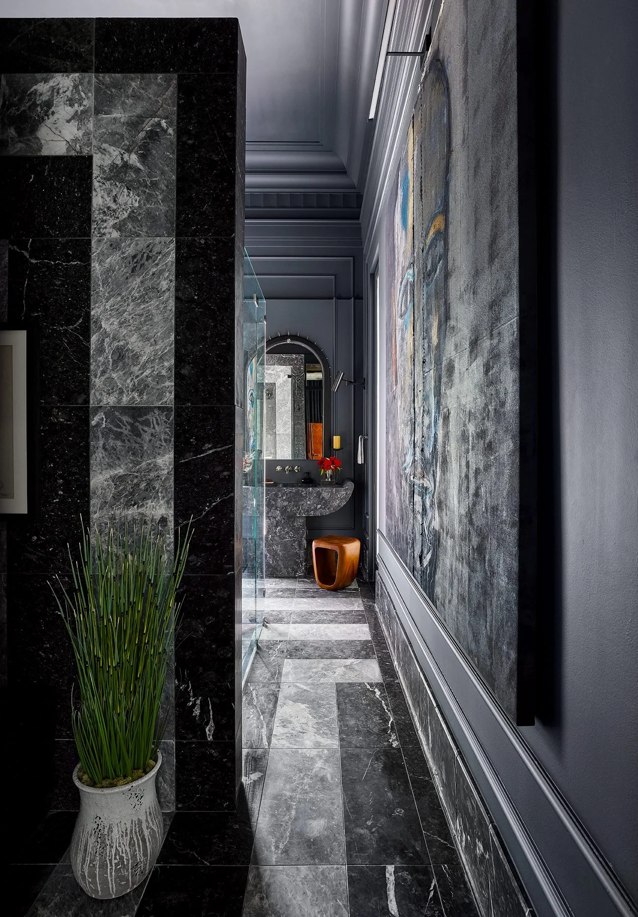 A modern interior space with dark marble features, a floor-to-ceiling mural or textured wall art, and a small vanity area with a curved mirror, a yellow candle, and a red flower arrangement. There is a potted green plant in a white vase in the foregr