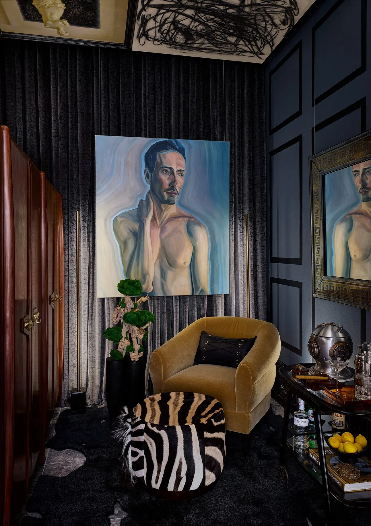 A modern living room corner with dark curtains, a large portrait of a shirtless man with dark hair, a yellow velvet armchair with a black pillow, a zebra-print ottoman, a small side table with various items, a plant in a black pot, and framed art on 