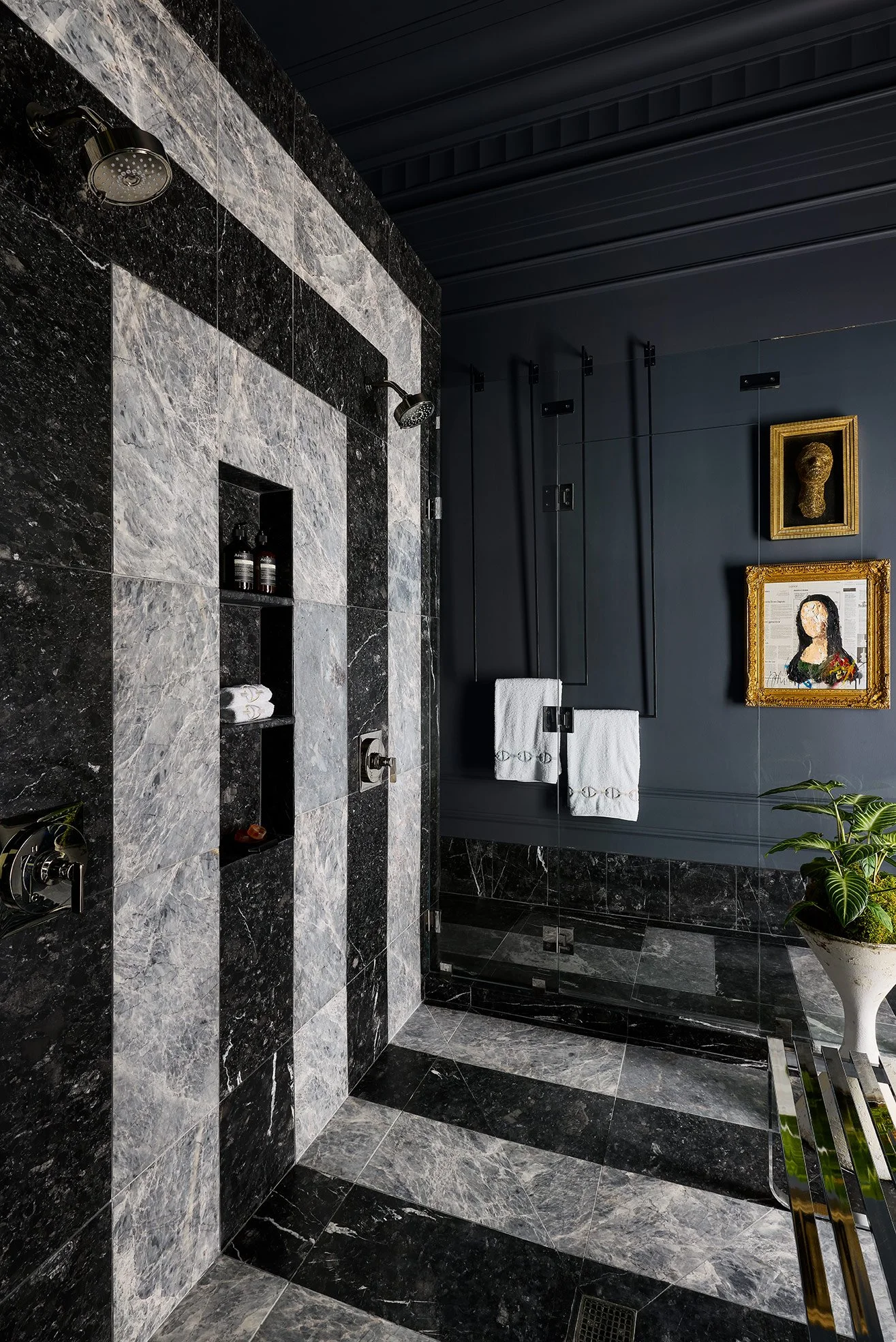 Modern bathroom with black and gray marble tiles, black paneled wall, framed art, and a glass shower enclosure.
