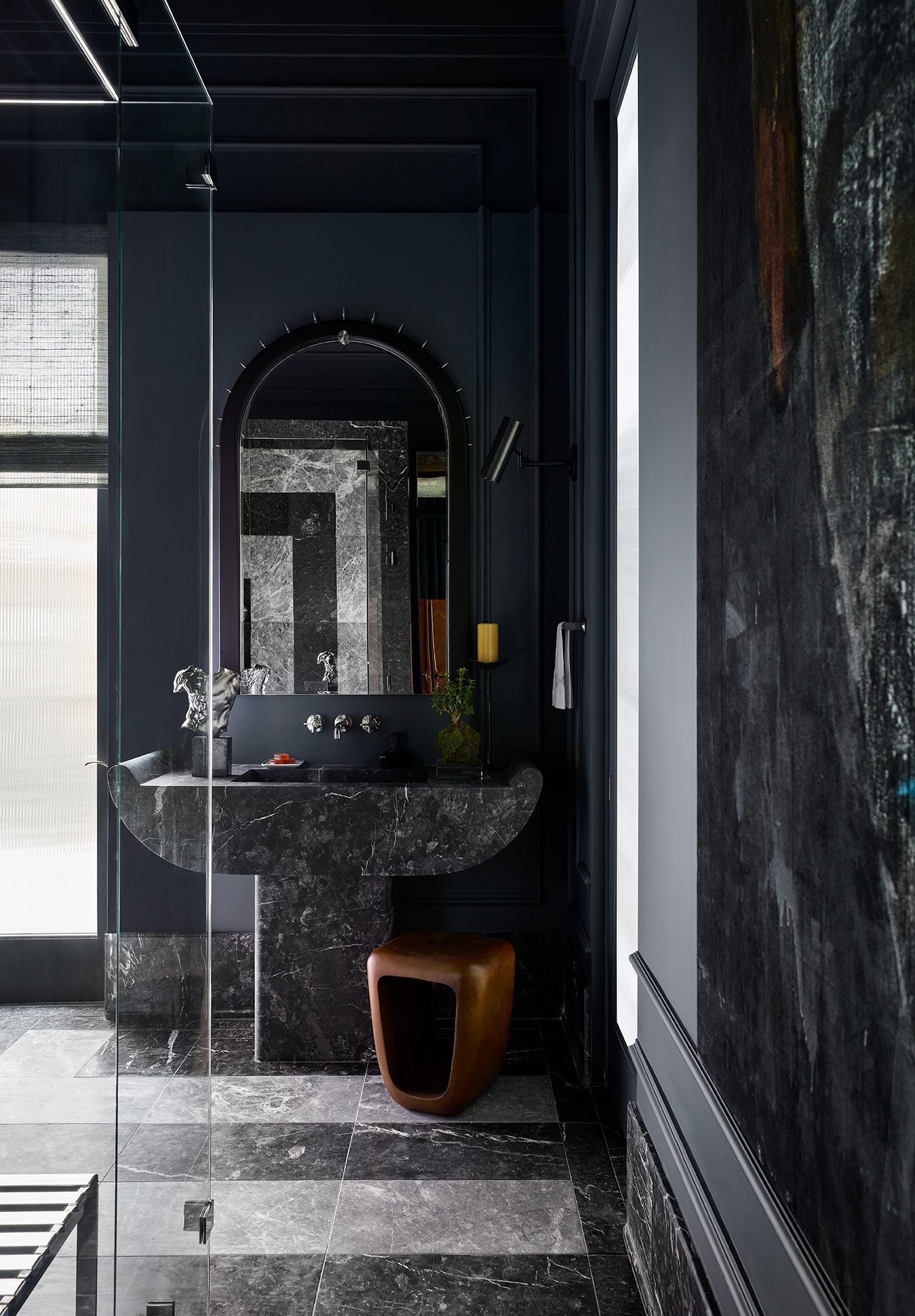 Modern bathroom with dark walls and marble countertops, featuring a mirror, a small plant, and a wooden stool.