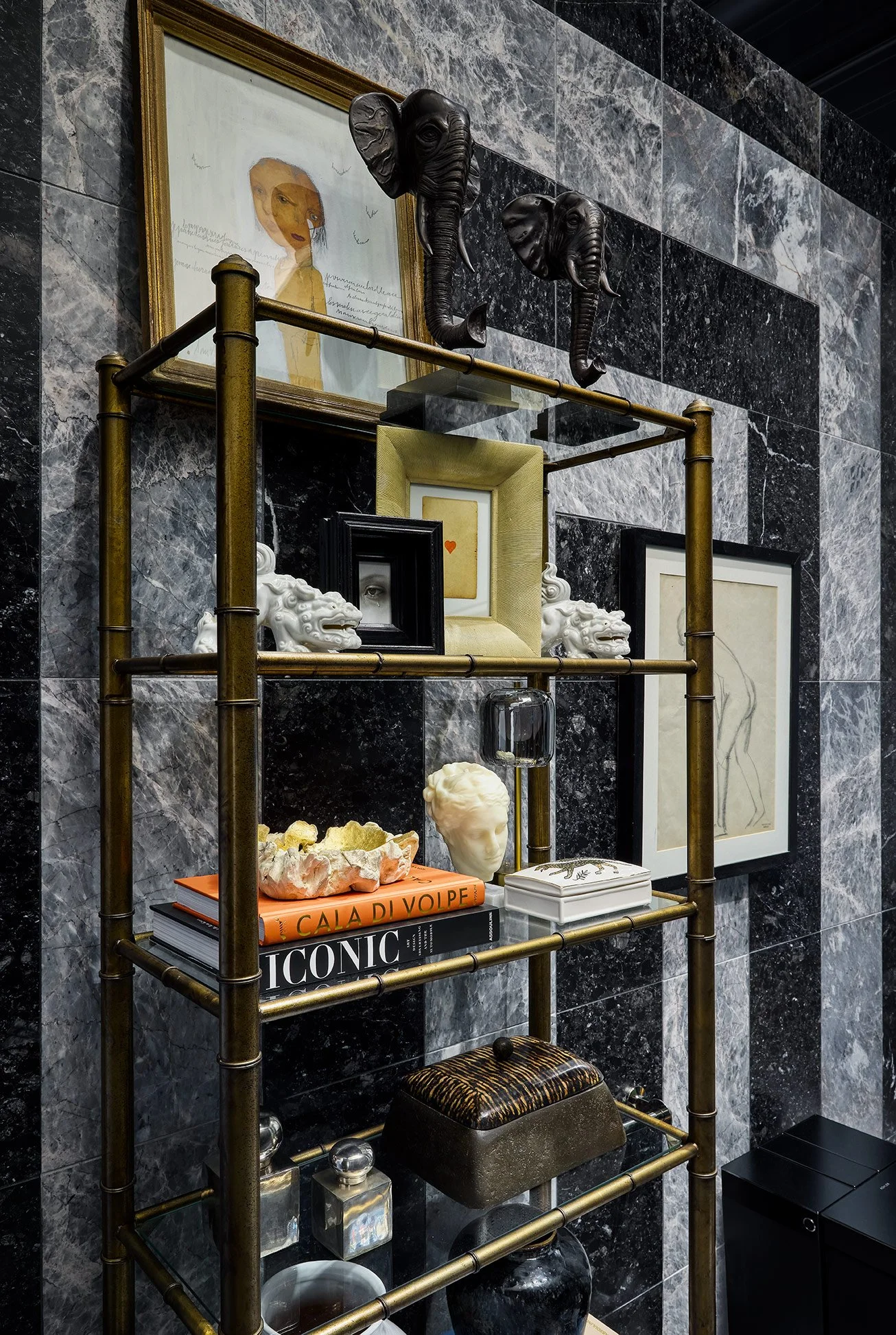 Decorative shelving unit against a gray marble wall holding art books, sculptures, framed artwork, and decorative items, with two elephant bust sculptures mounted on the wall above.