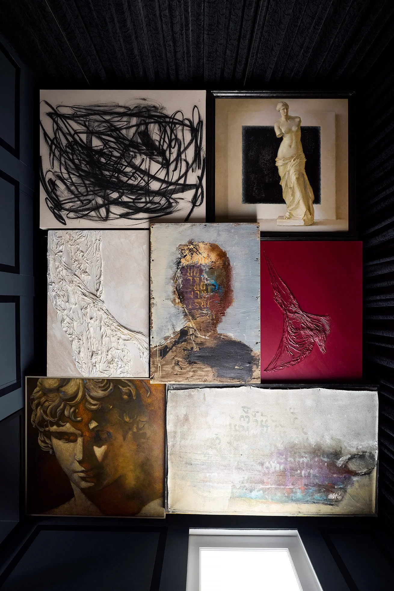 A collection of various art paintings displayed on a dark wall in a gallery. The artworks include abstract expression, classical sculpture, textured portrait, and mixed media pieces.