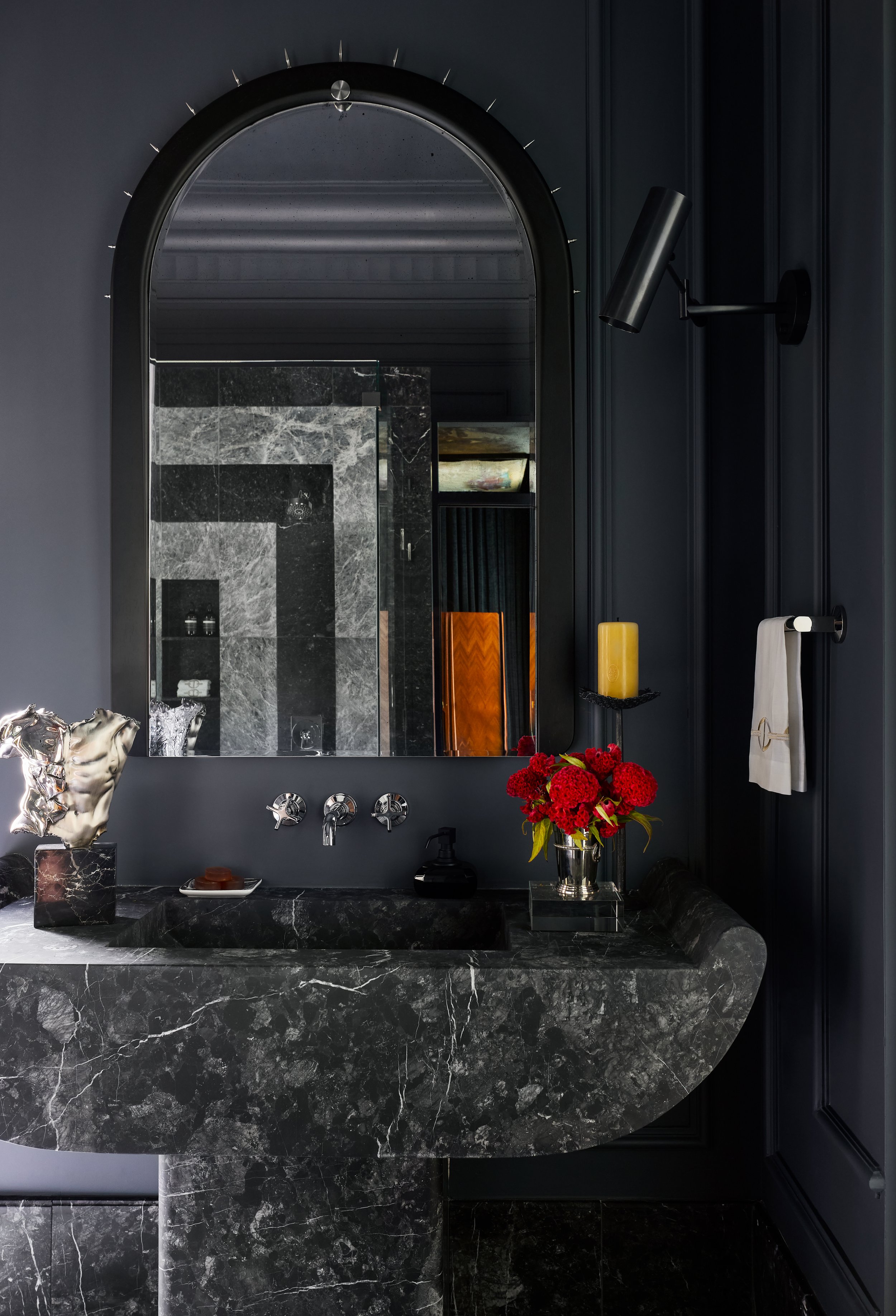 Bathroom sink vanity with a dark marble countertop, a large mirror, a black wall-mounted light, a tray of soap, a vase with red flowers, and a soap dispenser.
