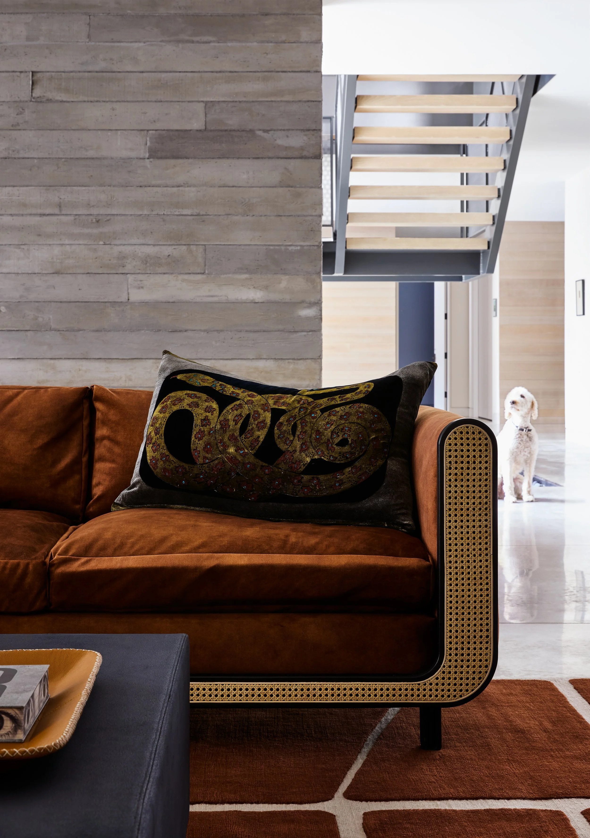 Modern living room with brown velvet sofa, decorative pillow, and area rug, featuring a staircase with light wood steps and metal railing in the background, and a small white dog standing near a door in the distance.