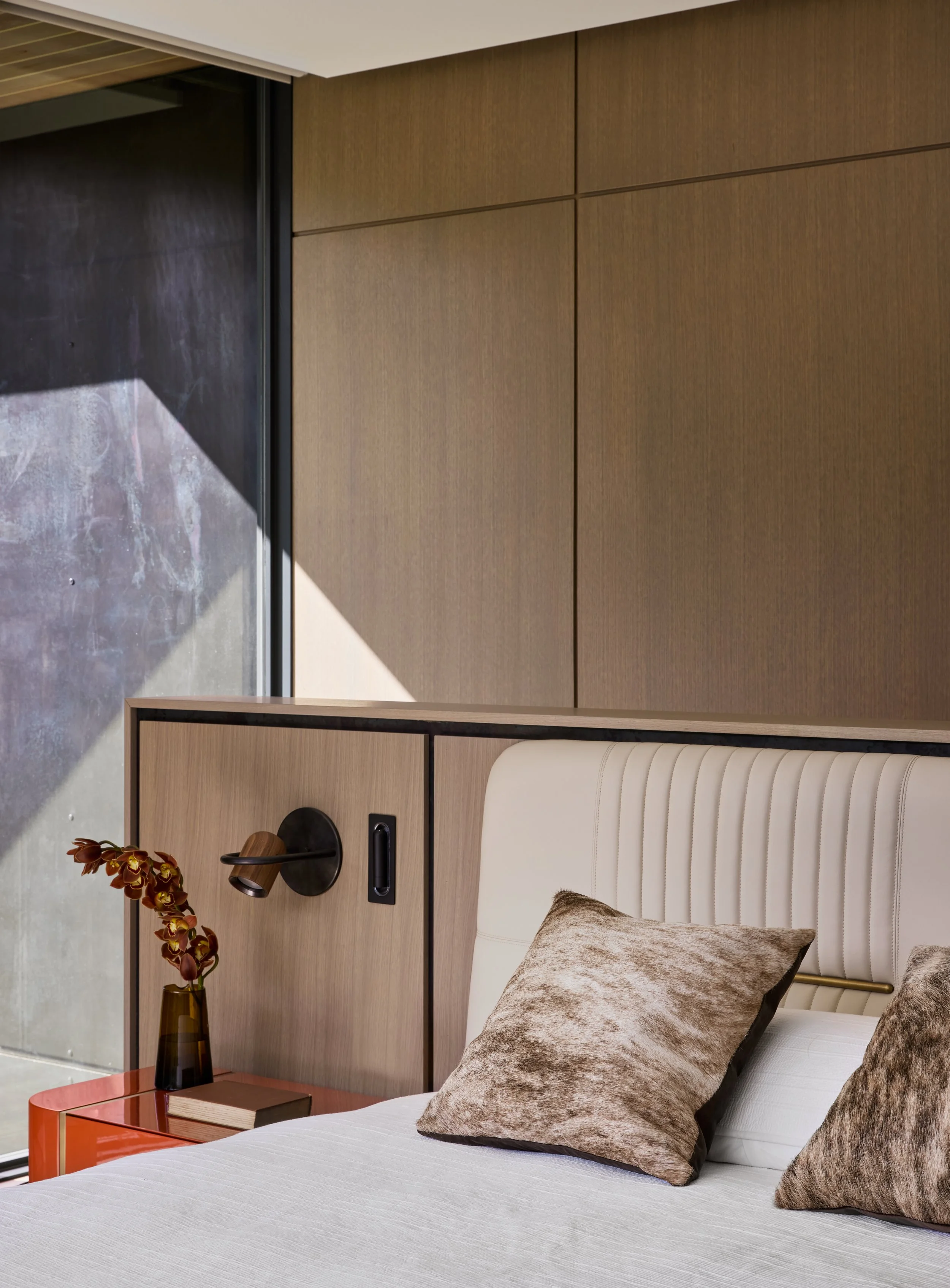 A modern bedroom with a white bed, brown and black textured pillows, a bedside table with a pink top, books, and a vase with brownish-red flowers. There's a wooden headboard, a wall-mounted black lamp, and a wooden panel wall. A large window with a b