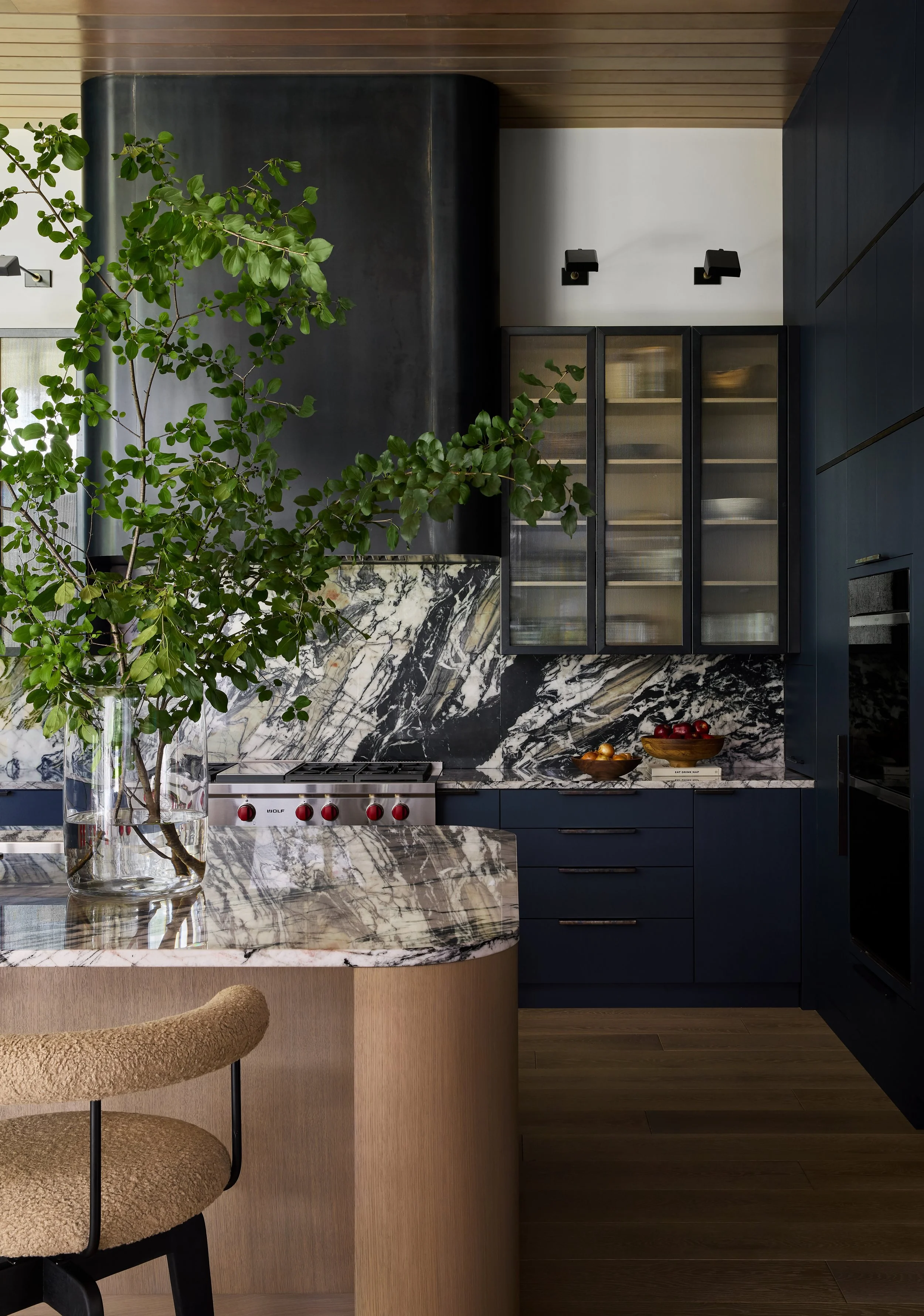 Modern kitchen with a marble island, blue cabinets, glass-fronted upper cabinets, and black wall-mounted lights. A green leafy plant is on the island.