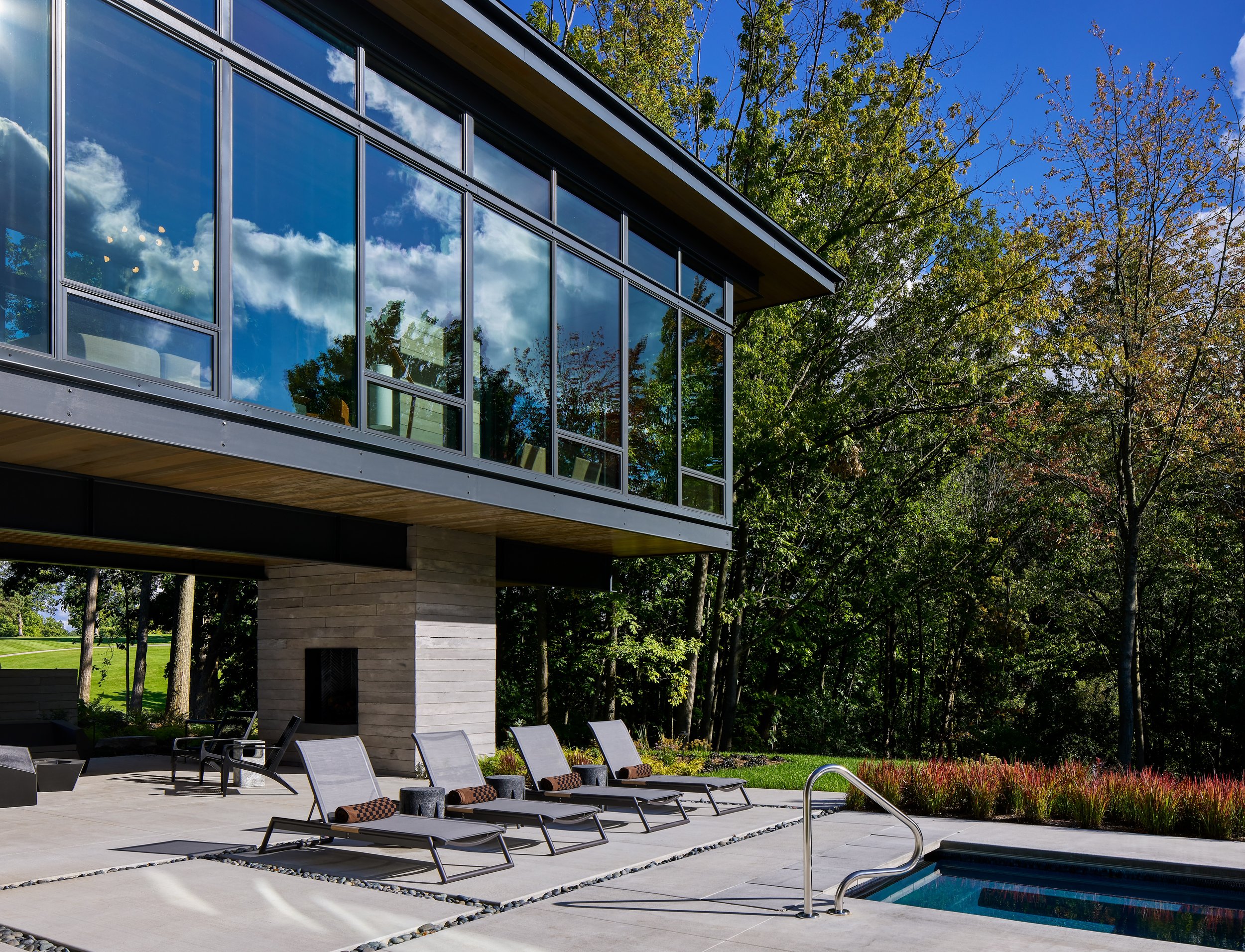 Modern house with large glass windows reflecting blue sky and clouds, surrounded by tall trees, with an outdoor pool and lounge chairs in the foreground.