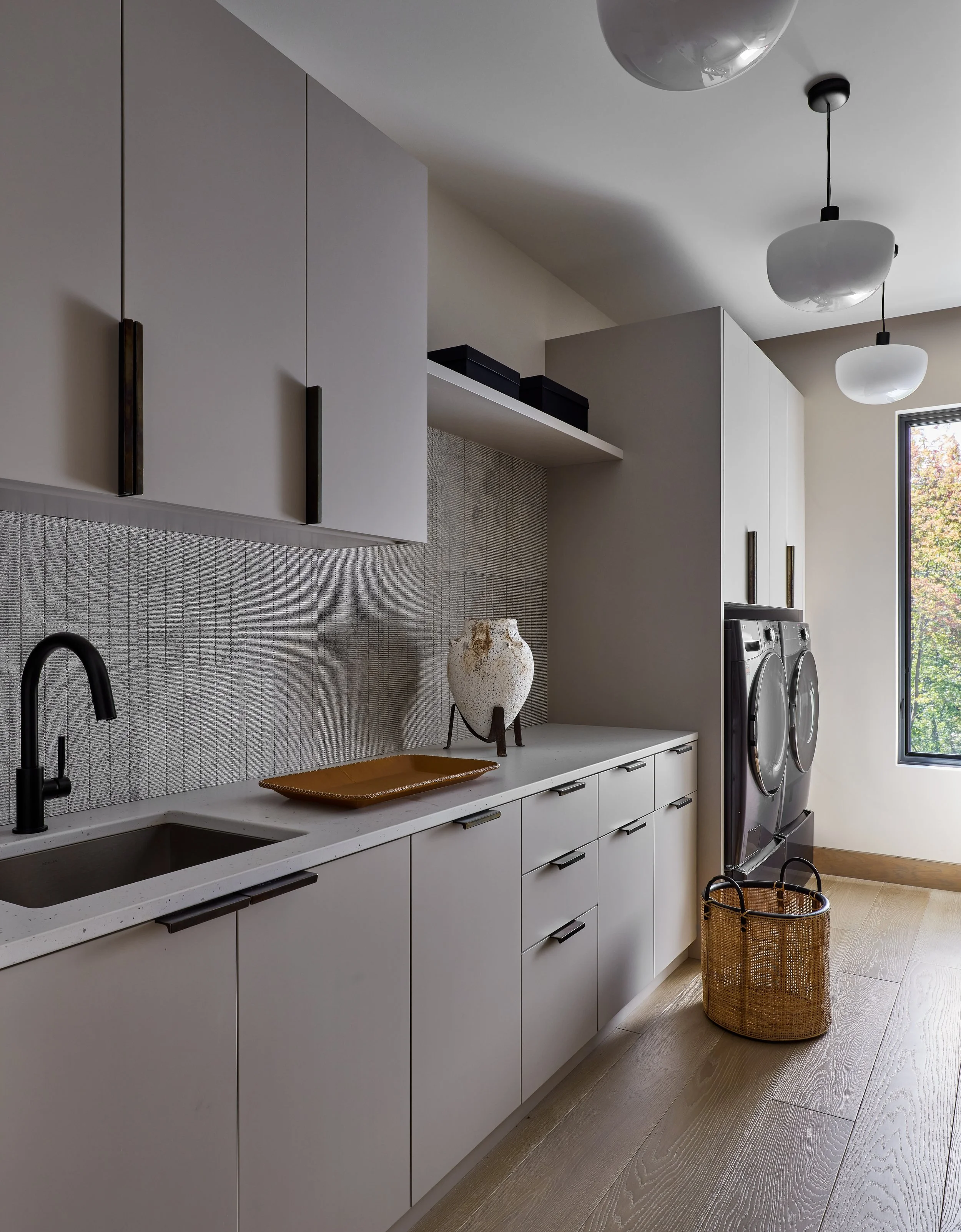 Modern laundry room with white cabinets, a black faucet, a textured backsplash, a decorative vase, a wicker laundry basket, and front-loading washer and dryer units near a window with a view of trees.