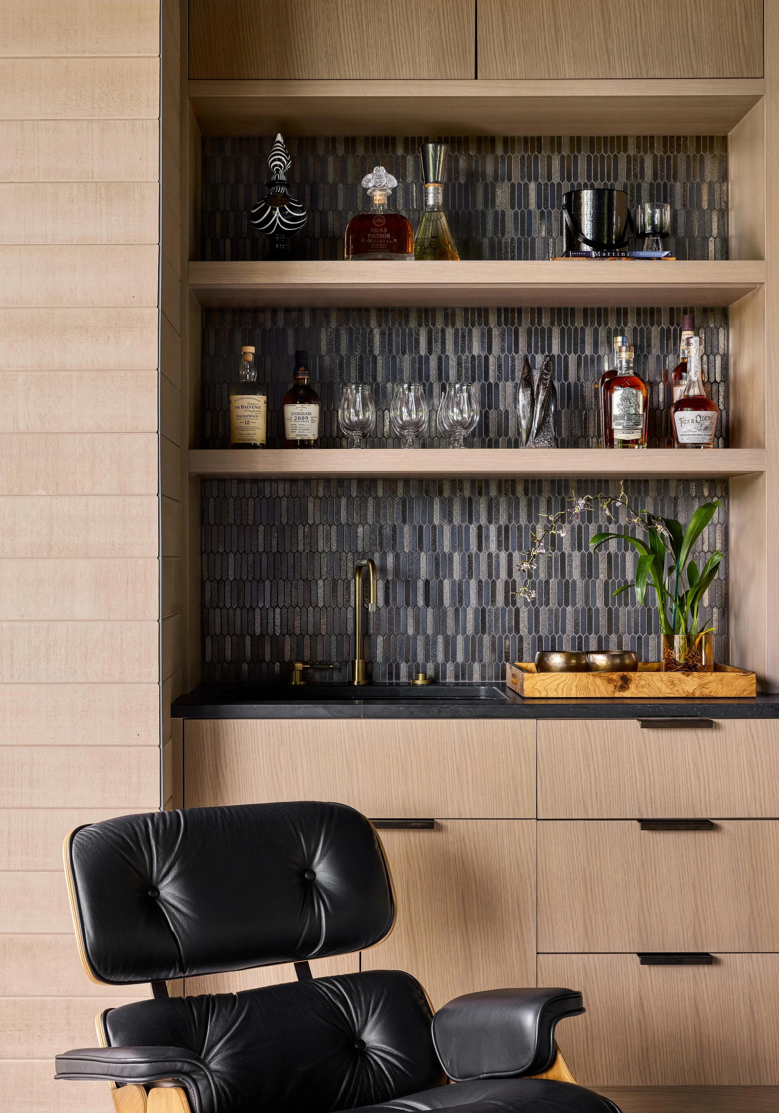 A modern kitchen with light wood cabinets, a black tile backsplash, wine bottles, glasses, and a plant on a wooden tray, with a black leather chair in front.