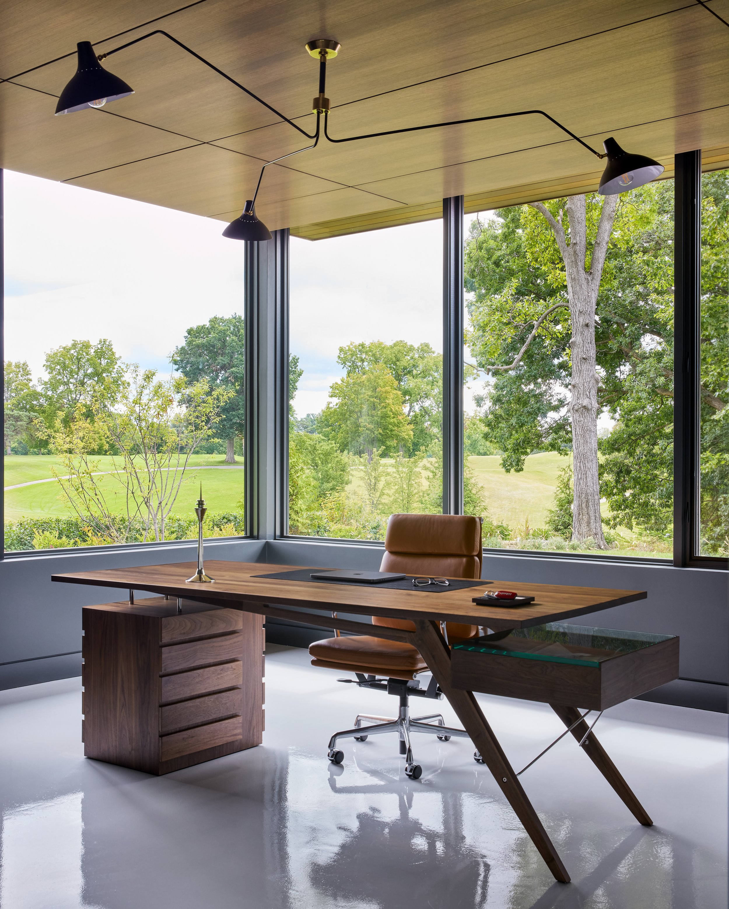 Modern office with large floor-to-ceiling windows showing a green outdoor landscape, a wooden desk with a chair, a laptop, a candle holder, glasses, and a minimalist ceiling light fixture.