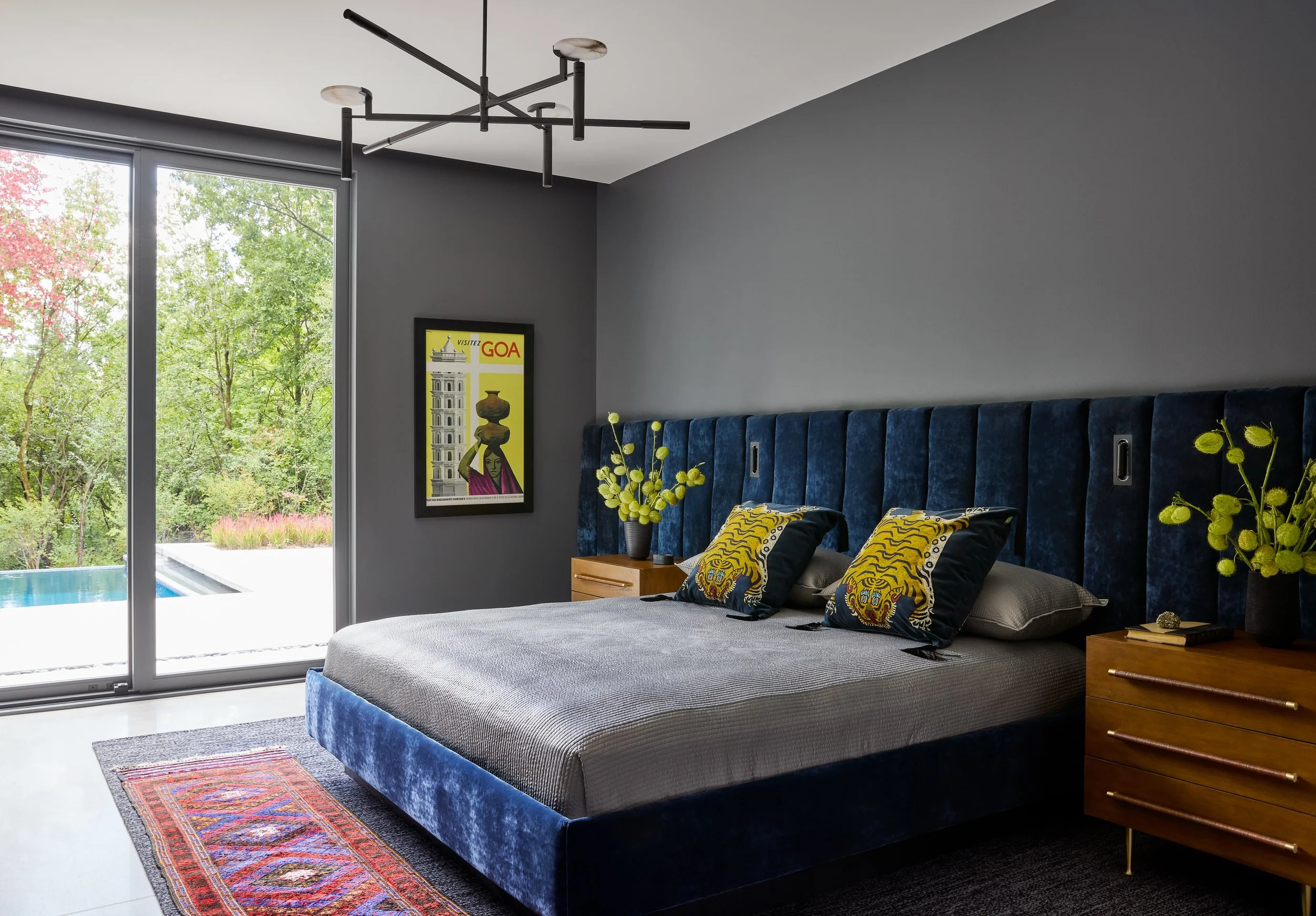 Modern bedroom with large window showing outdoor greenery, gray walls, a navy blue upholstered bed with tiger print pillows, yellow accent pillows, a small wooden nightstand with flowers, a framed poster of Goa in India, and a contemporary ceiling li