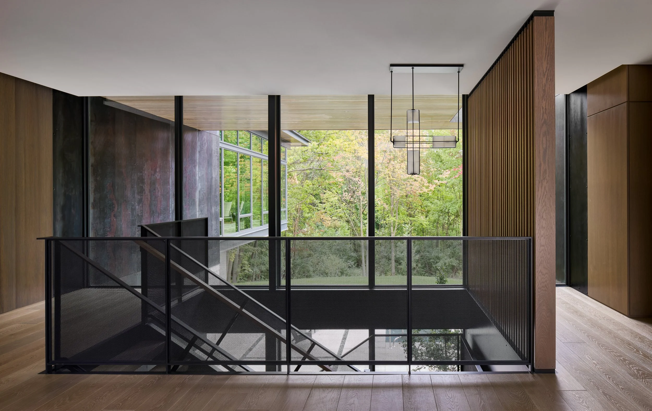 Interior view of a modern building with large floor-to-ceiling windows showing trees outside, wooden accents, and hanging ceiling lights above a staircase enclosed with a black metal railing.
