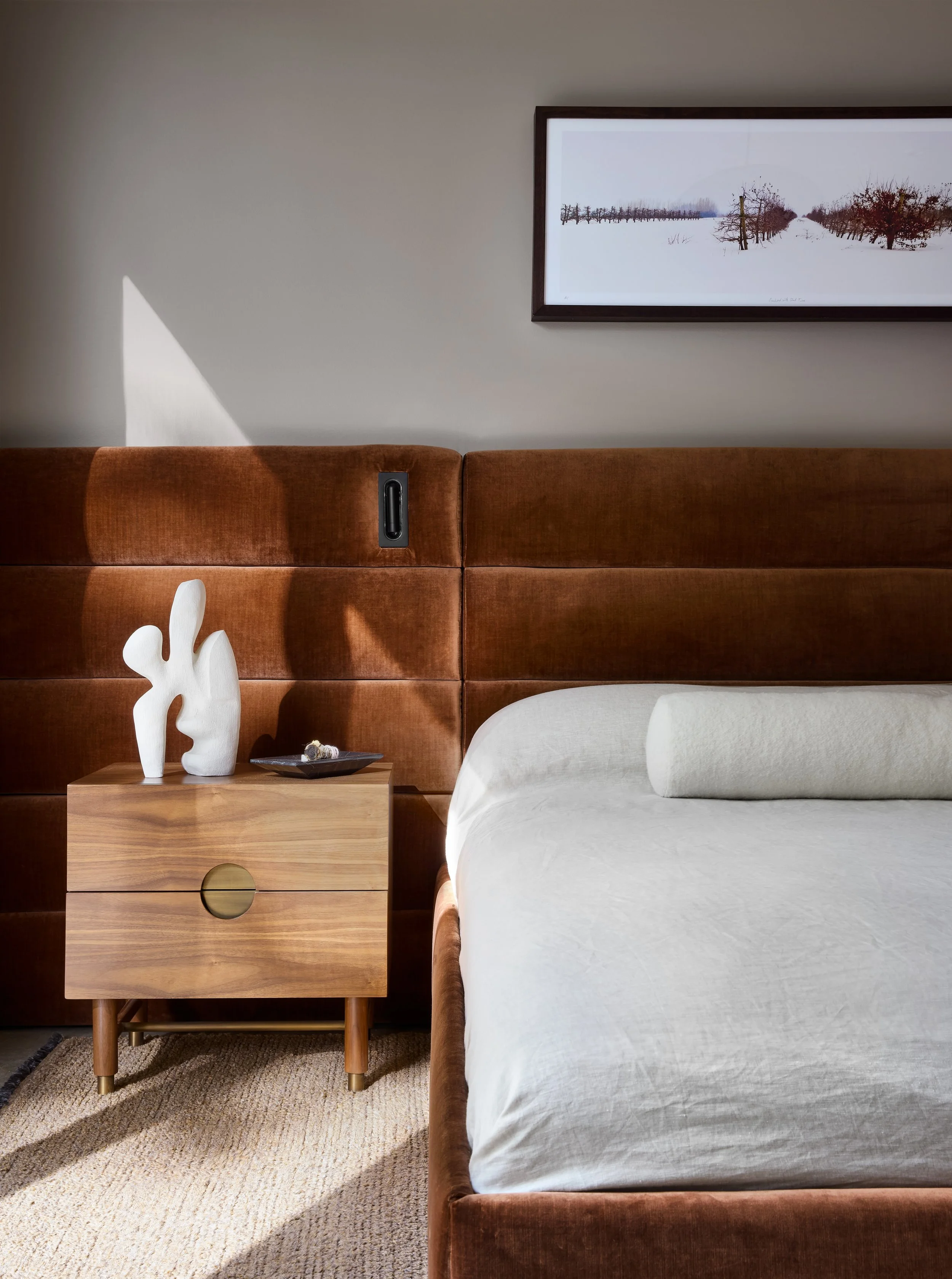 Interior of a bedroom showing a bed with a white duvet, a tan upholstered headboard, a wooden nightstand with white decorative objects, a framed winter landscape painting, and a carpeted floor.