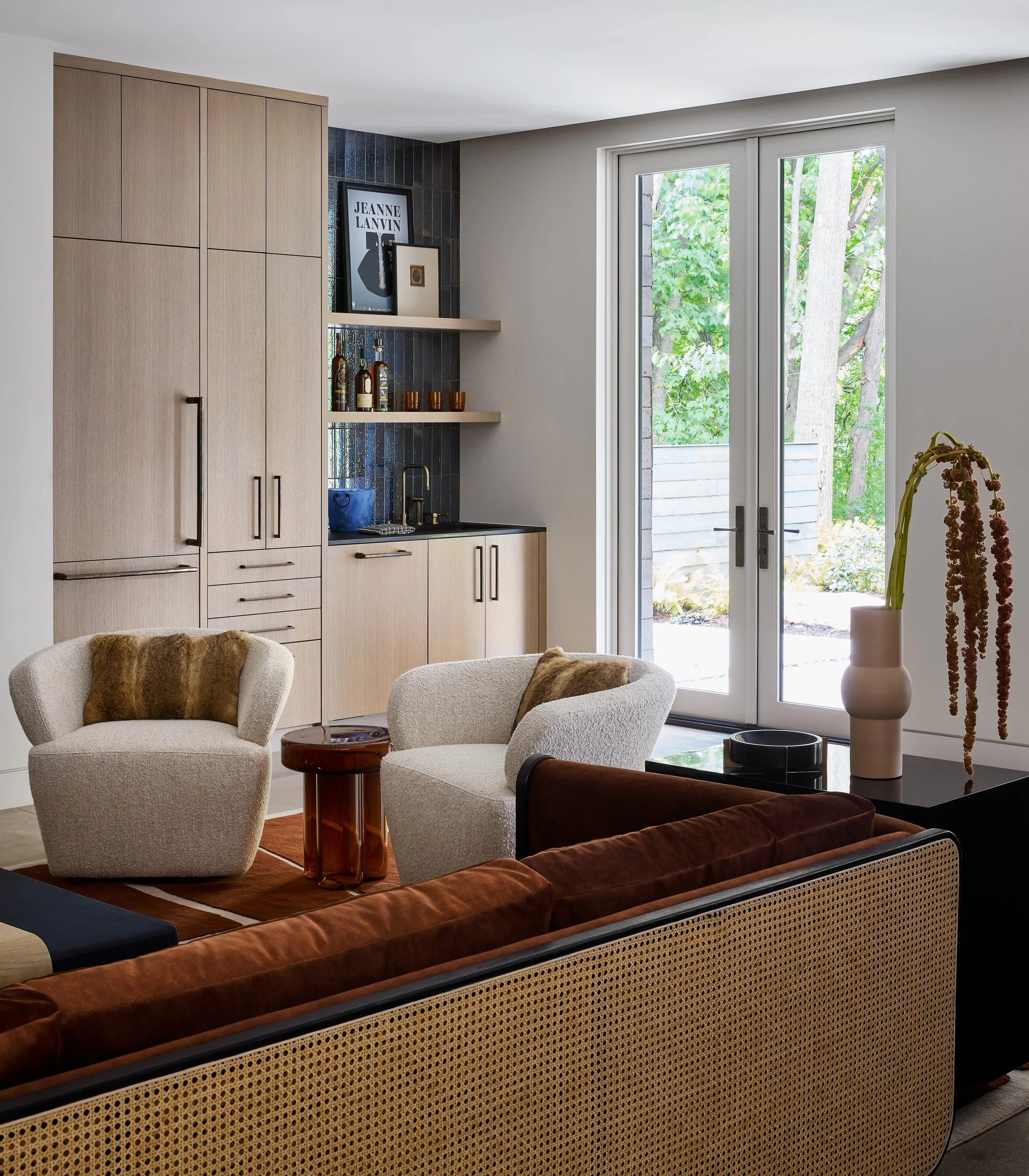 Living room with beige armchairs, a black and brown sofa, a wooden coffee table, a built-in kitchen cabinet, decorative bottles, artwork, and large glass door leading outside with greenery visible