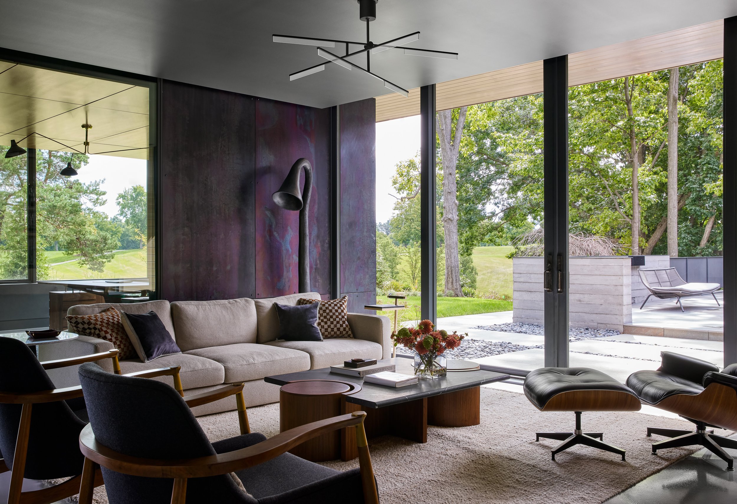 Modern living room with large floor-to-ceiling windows showing green trees outside, beige couch with patterned pillows, black and wooden chairs, coffee table with books and flowers, and an outdoor patio with lounge chairs.