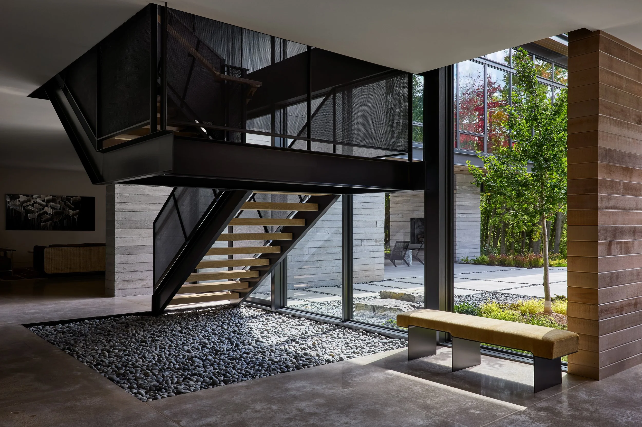 Modern interior with a black metal staircase, large glass windows showing trees and outdoor landscape, and a wooden wall on the right.
