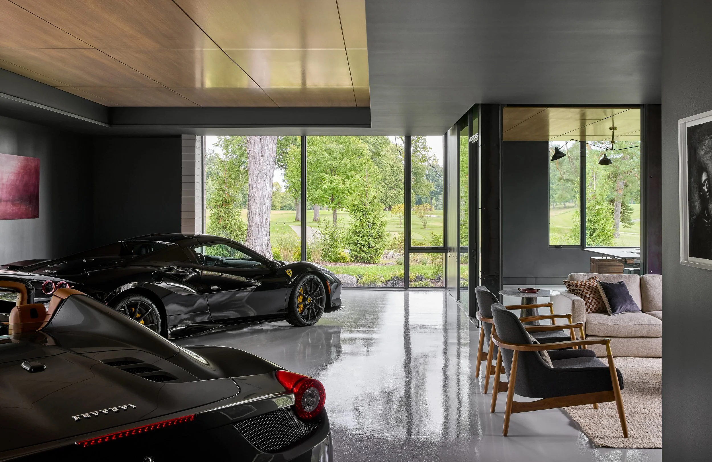Interior of a modern garage with a black Ferrari sports car and a silver Ferrari, viewing the outdoors through large glass windows showing trees and greenery.