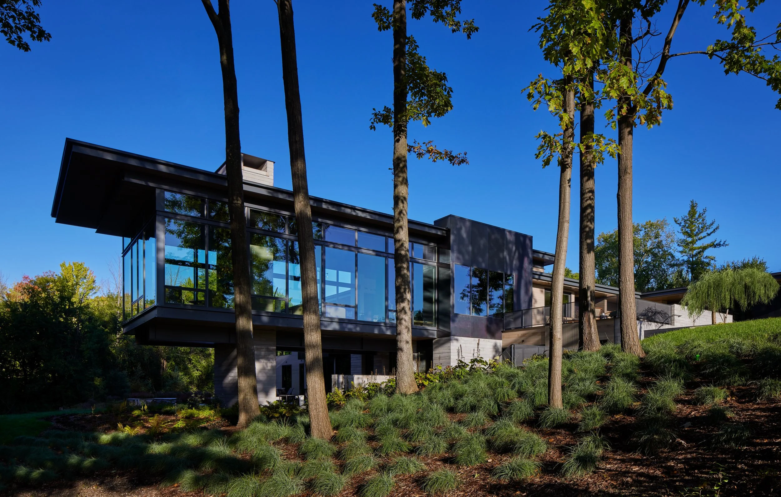 Modern house with large glass windows surrounded by trees, situated on a hillside under a clear blue sky.