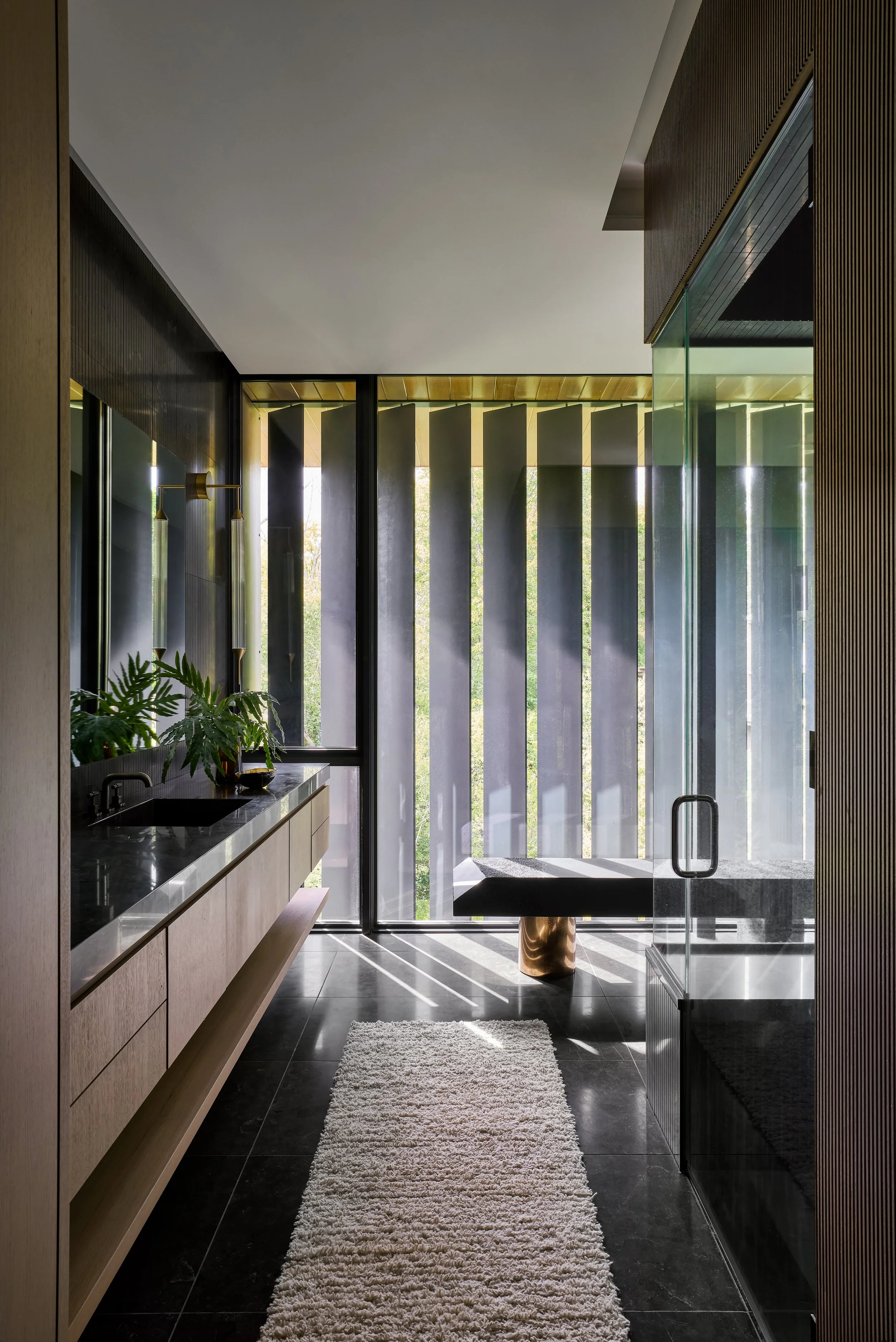 Modern interior bathroom with black tile floor, beige wooden cabinets below a black speckled countertop, decorative lighting, and large windows with vertical slats and natural sunlight. There is a beige shaggy rug on the floor and a green plant near 