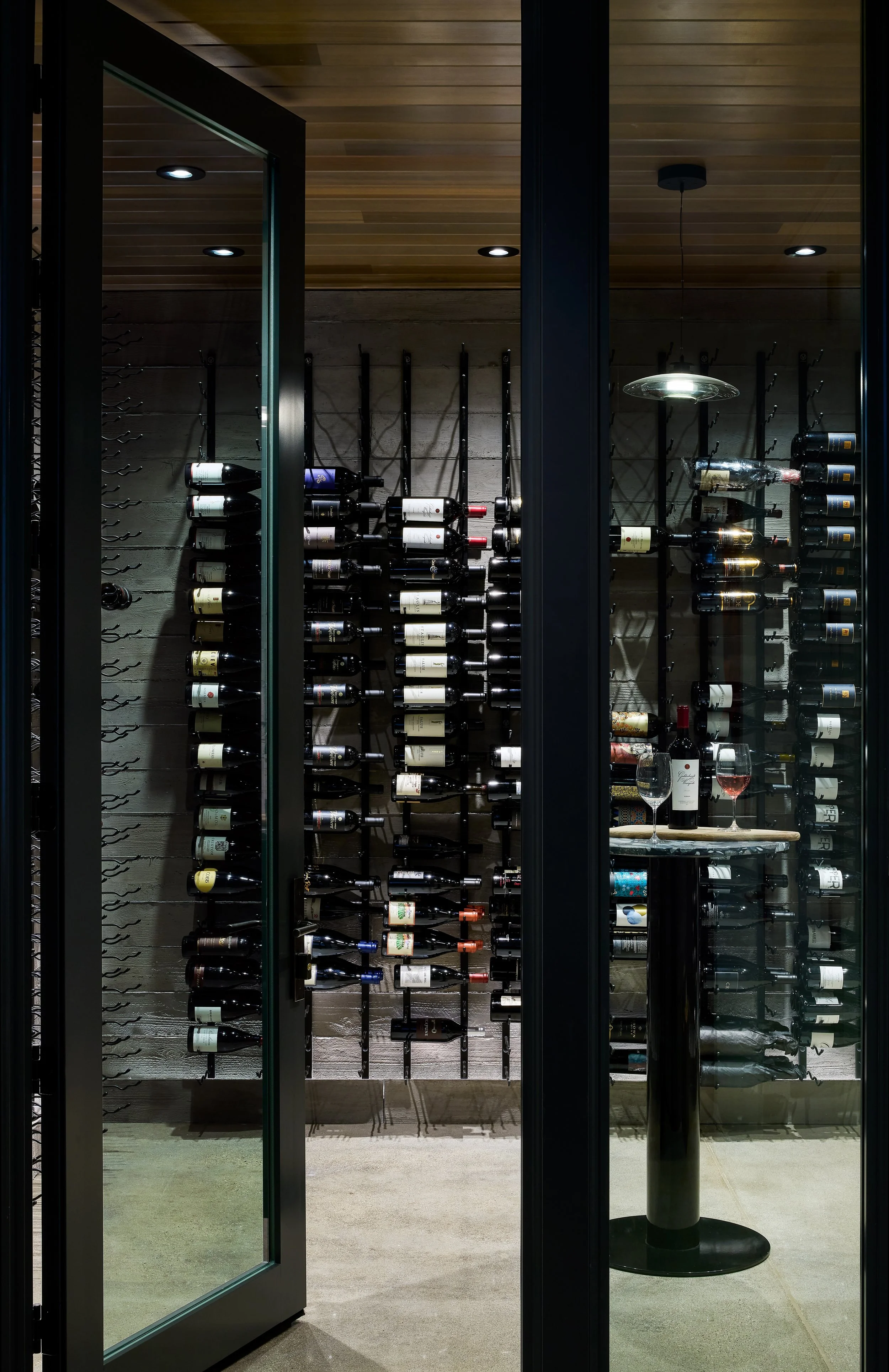 A wine cellar with wine bottles stored horizontally on wall-mounted racks behind a glass door, and a round table with wine bottles and glasses in front of the shelves.