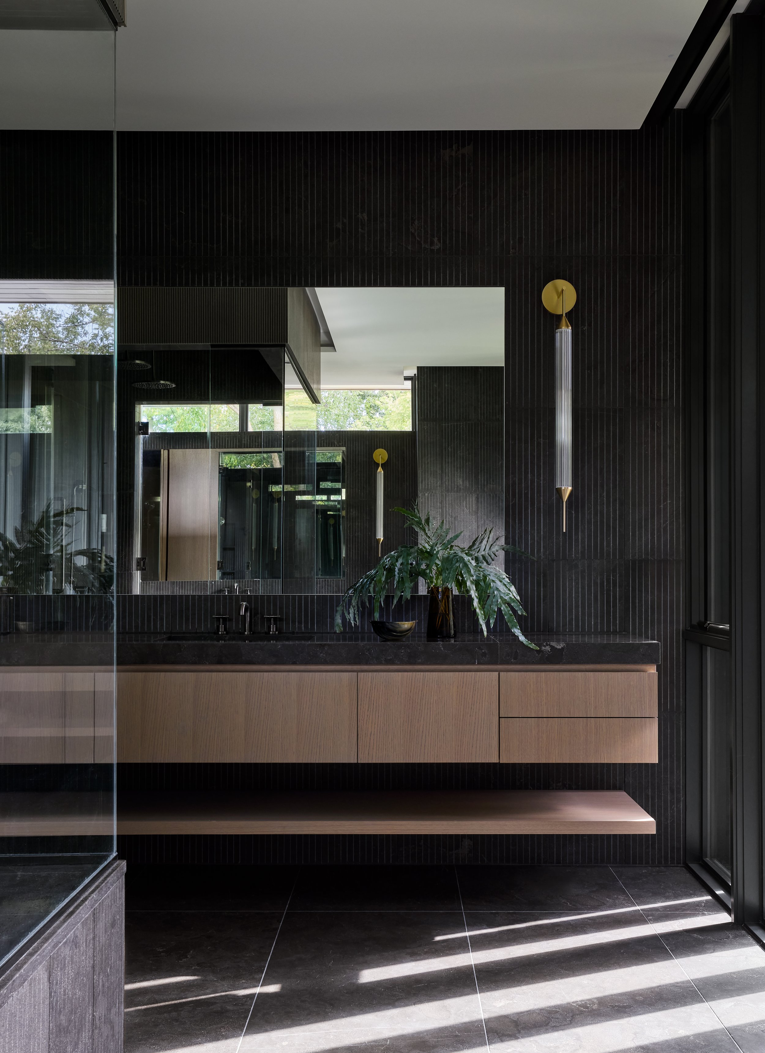 Modern bathroom with black textured tiles, a large mirror, a black countertop with a plant, modern lighting fixtures, and sunlight streaming through a glass door.