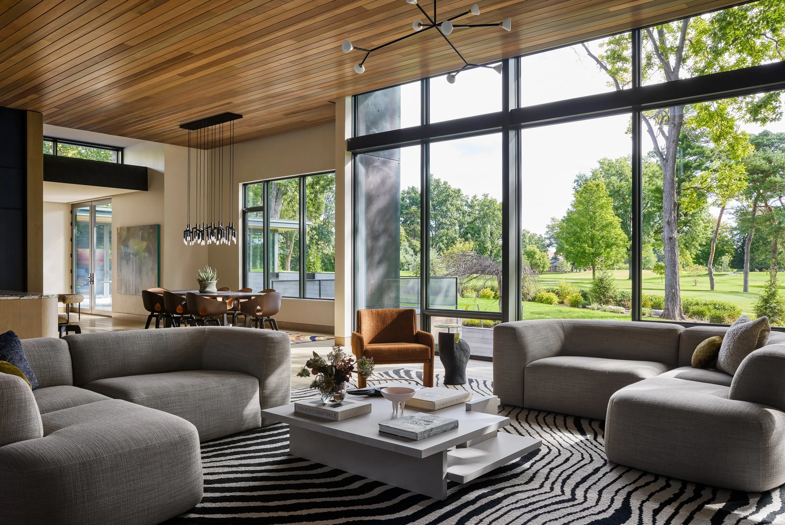 Modern living room with large floor-to-ceiling windows showing green trees outside, gray sofas, a white coffee table with books and decorative items, a black and white striped rug, and a wooden ceiling with contemporary light fixtures.