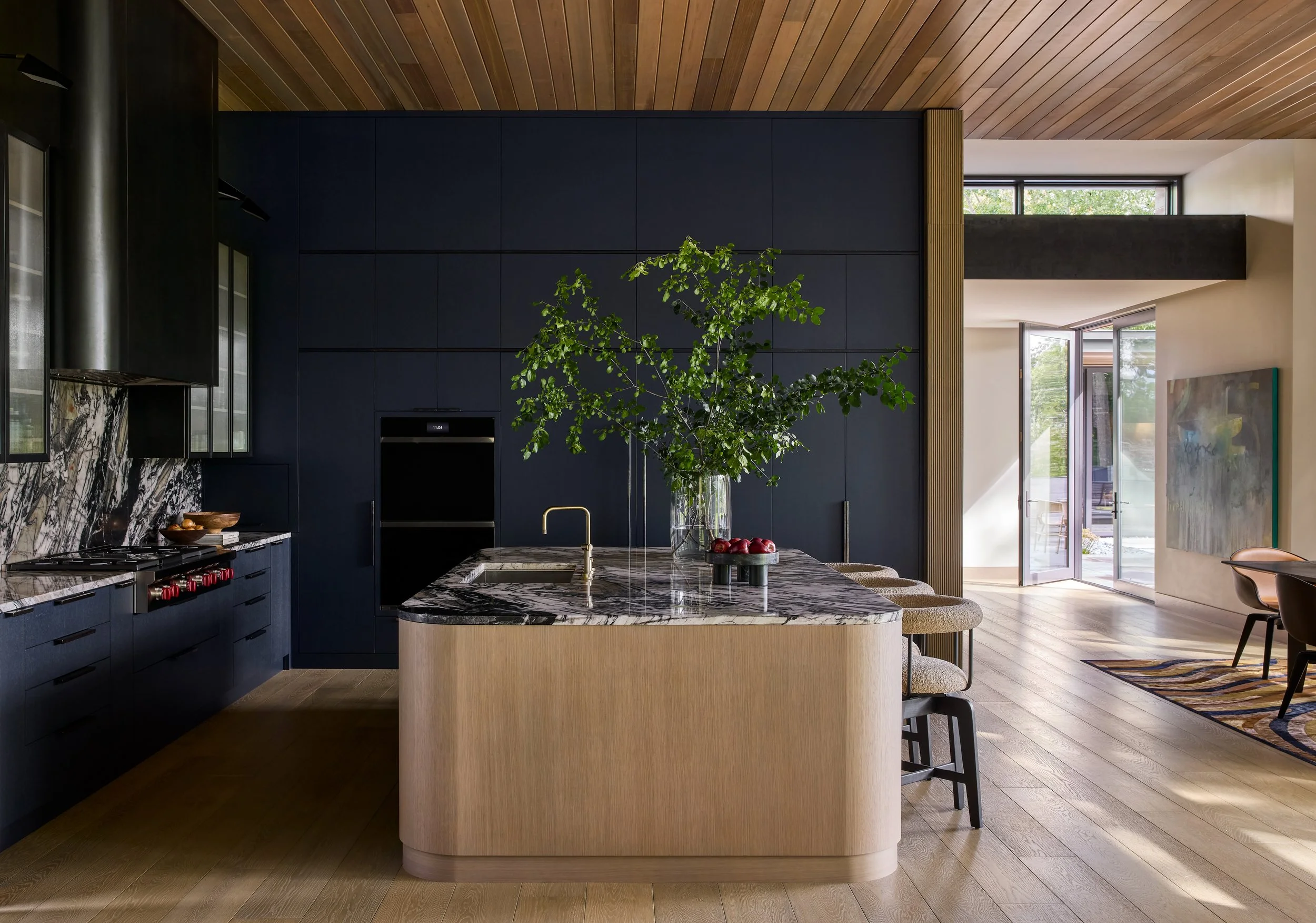 Modern kitchen with blue cabinets, marble countertops, a central island with a vase of green leafy plant, and wooden ceiling. Open sliding door reveals outdoor patio.