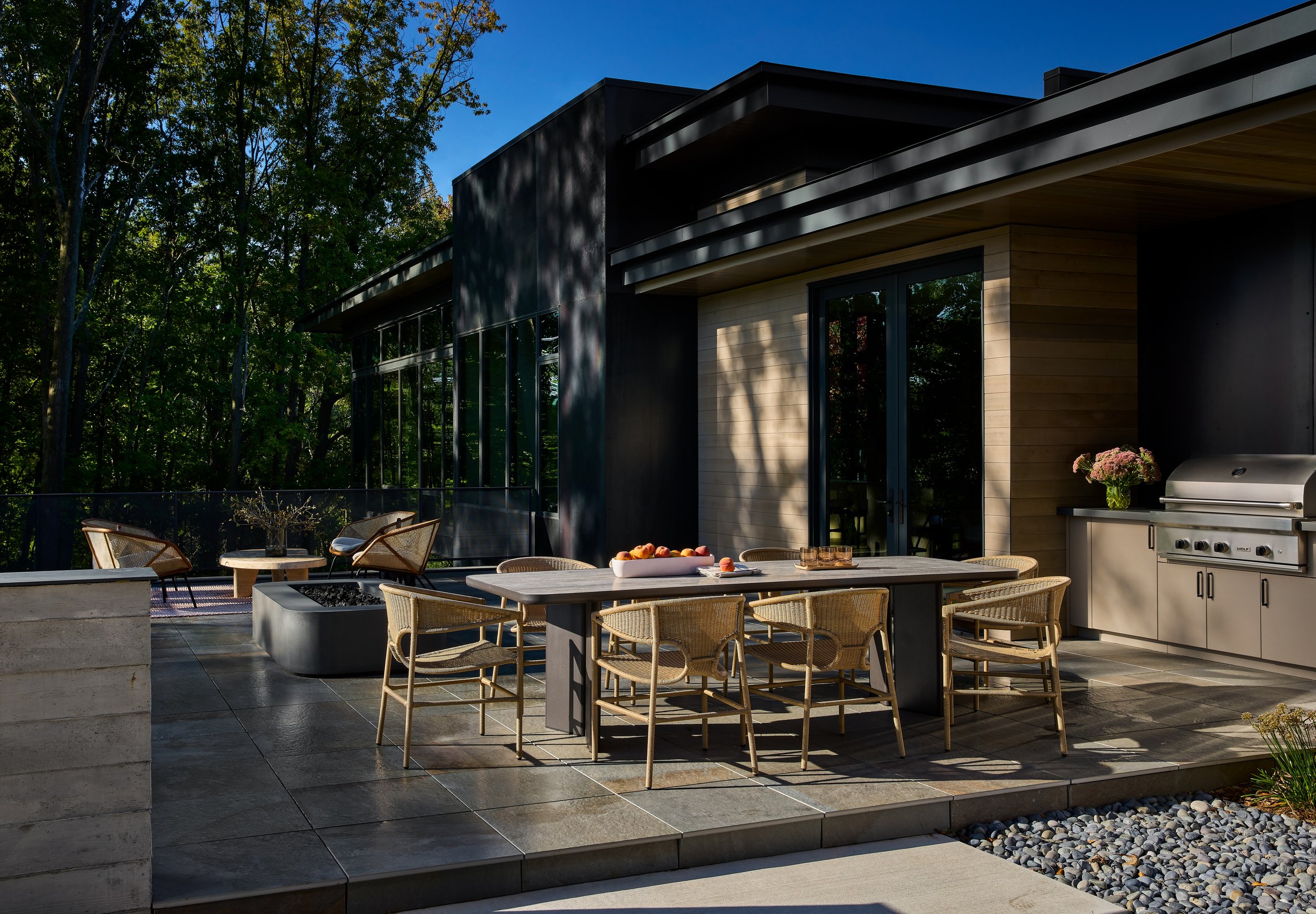 Modern house patio with outdoor dining area, wooden table, woven chairs, barbecue grill, potted flowers, and surrounding trees under a clear blue sky.